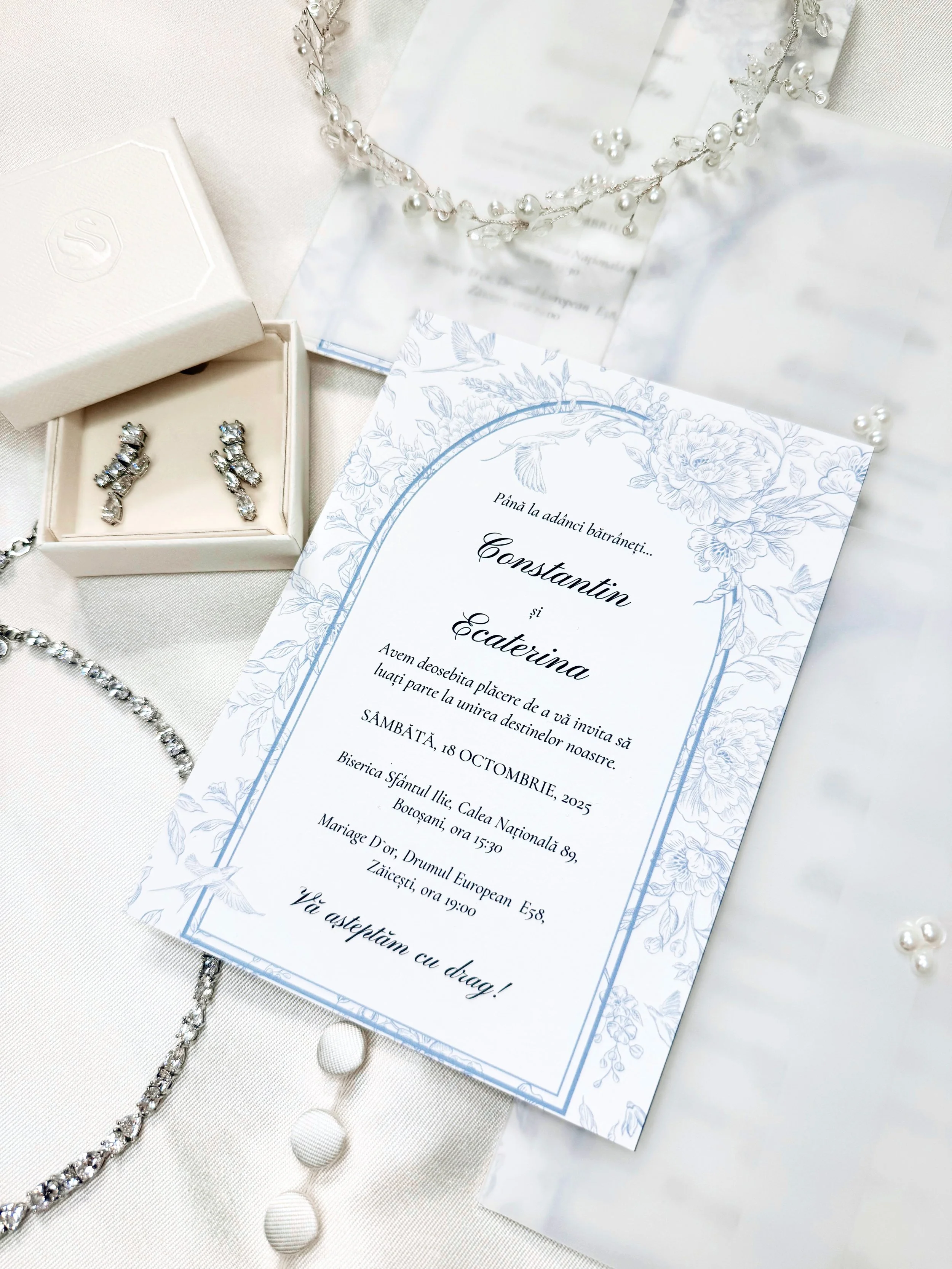 Wedding invitation card with blue floral design, silver jewelry including earrings and necklaces, a pearl headpiece, and white fabric background.