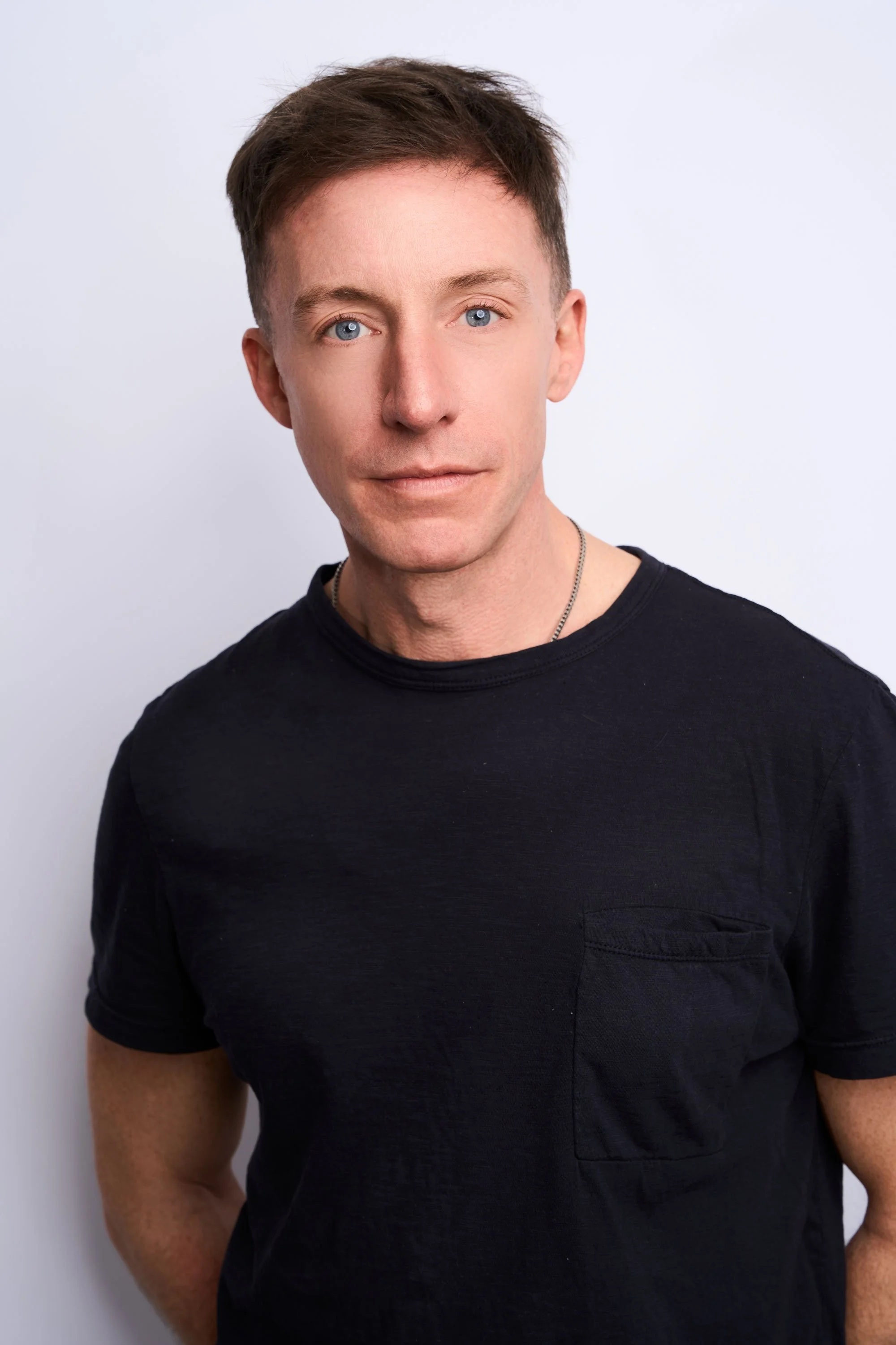 A young man with short brown hair and blue eyes, wearing a black t-shirt with a small pocket and a silver chain necklace, standing against a plain white background.