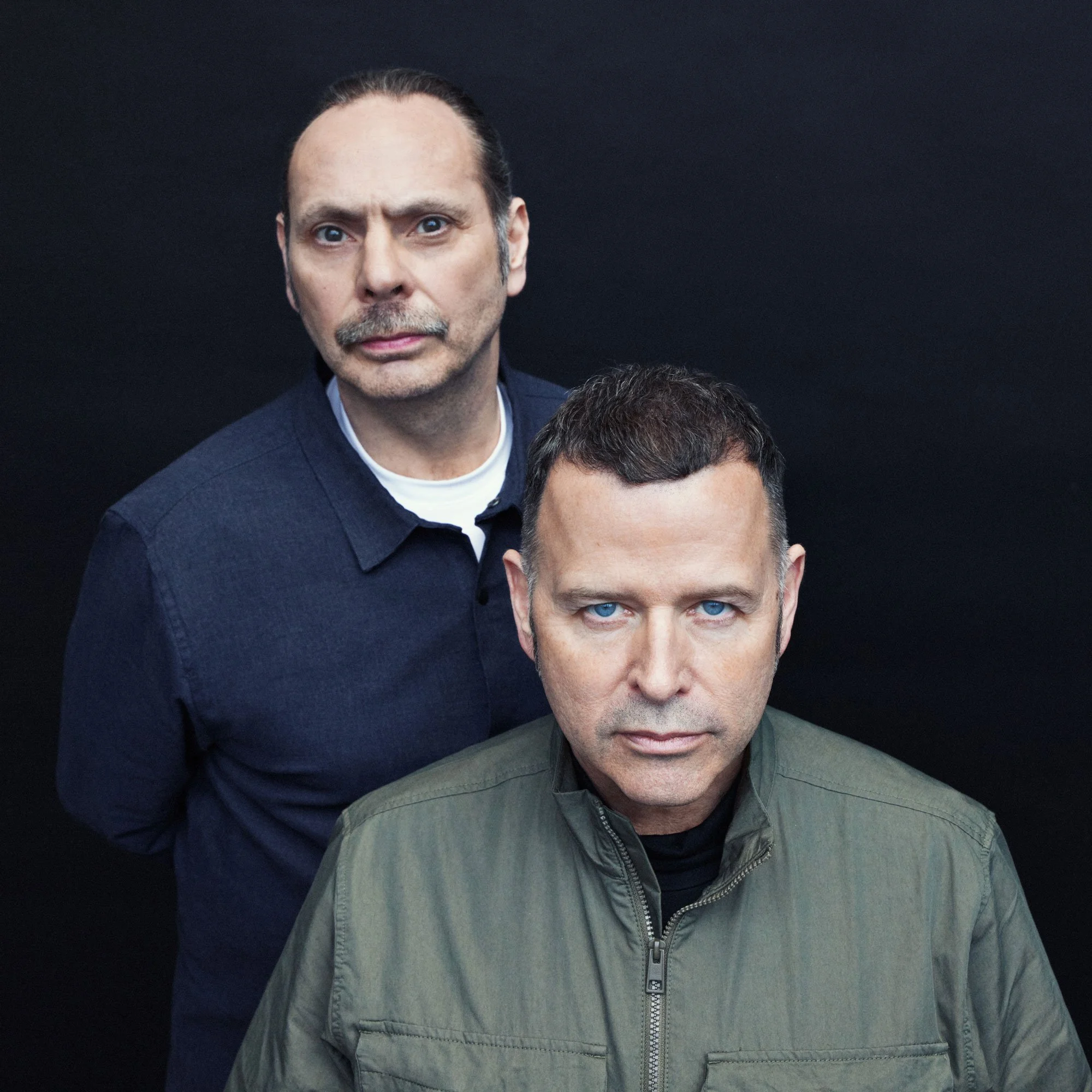 Two men with short dark hair and blue eyes posing against a black background, one standing behind the other. One man wears a dark blue shirt, and the other wears an olive green jacket.