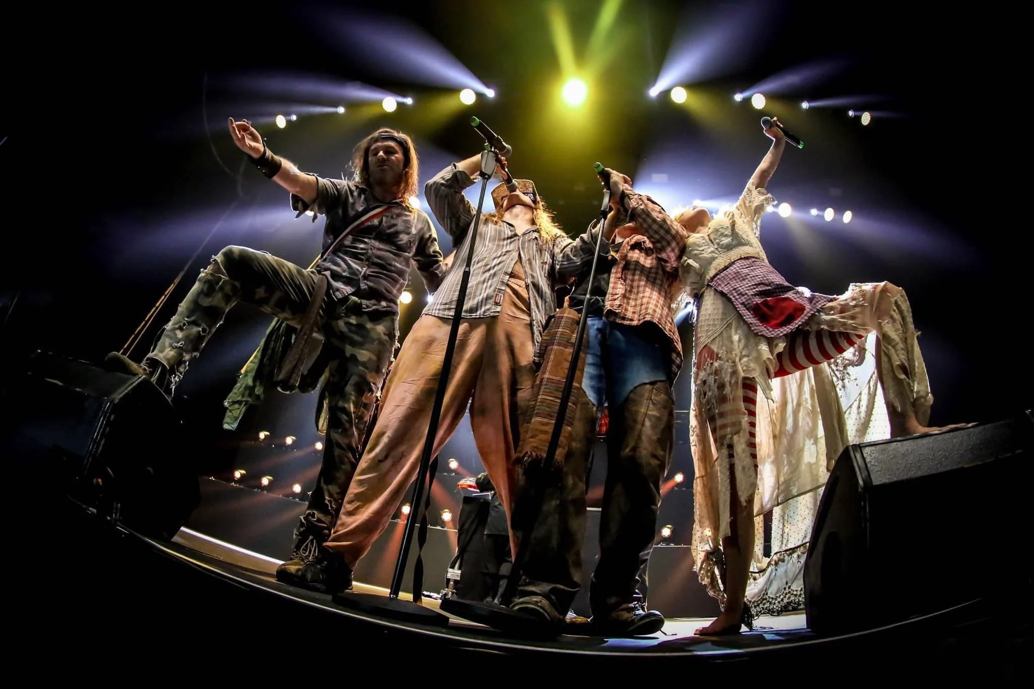 A group of five performers on stage dressed in eclectic, vintage-style clothing, singing into microphones with colorful lighting and stage lights above them.
