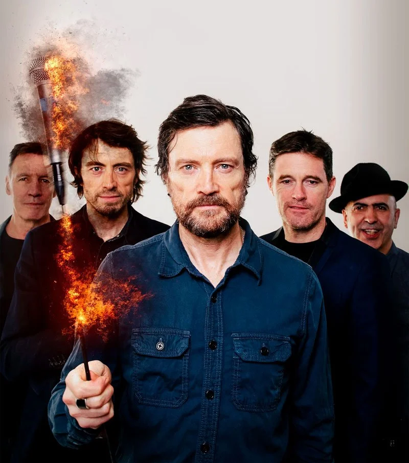 Five men standing in front of a plain background, with the man in the front holding a lit sparkler that appears to be on fire.