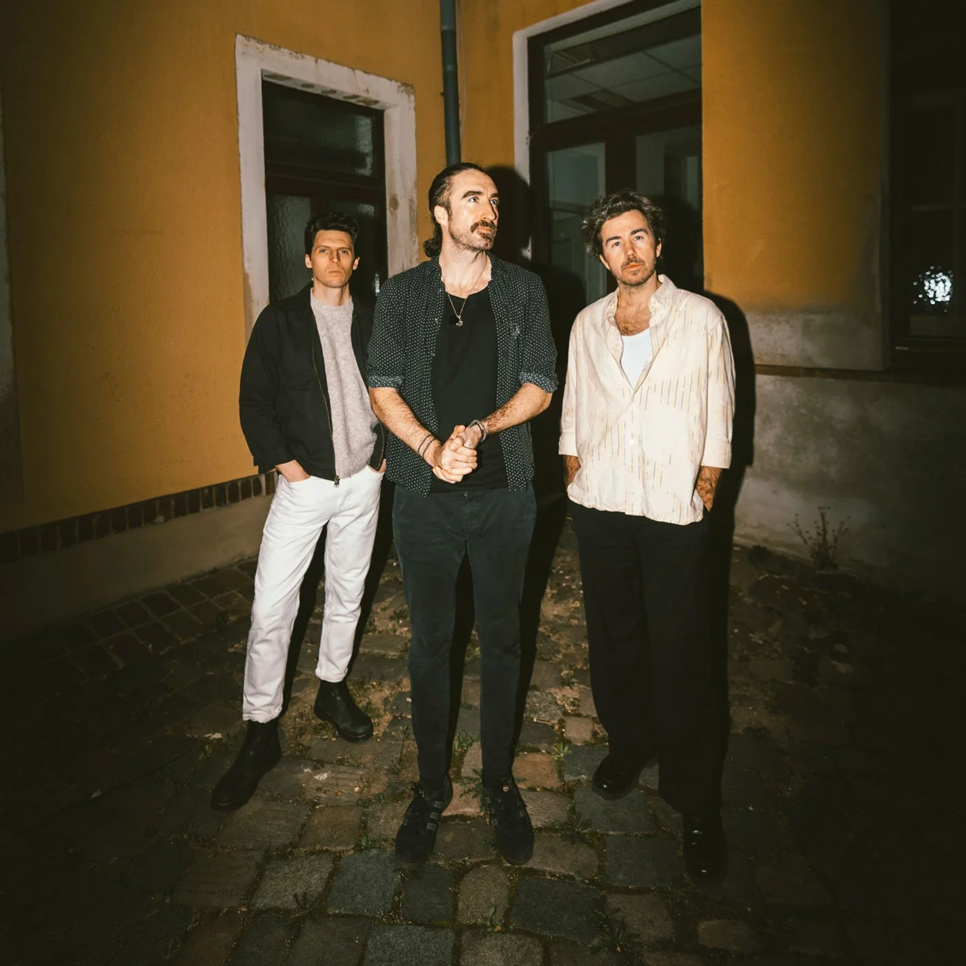 Three men standing outside at night on a cobblestone street, with yellow and gray buildings in the background.