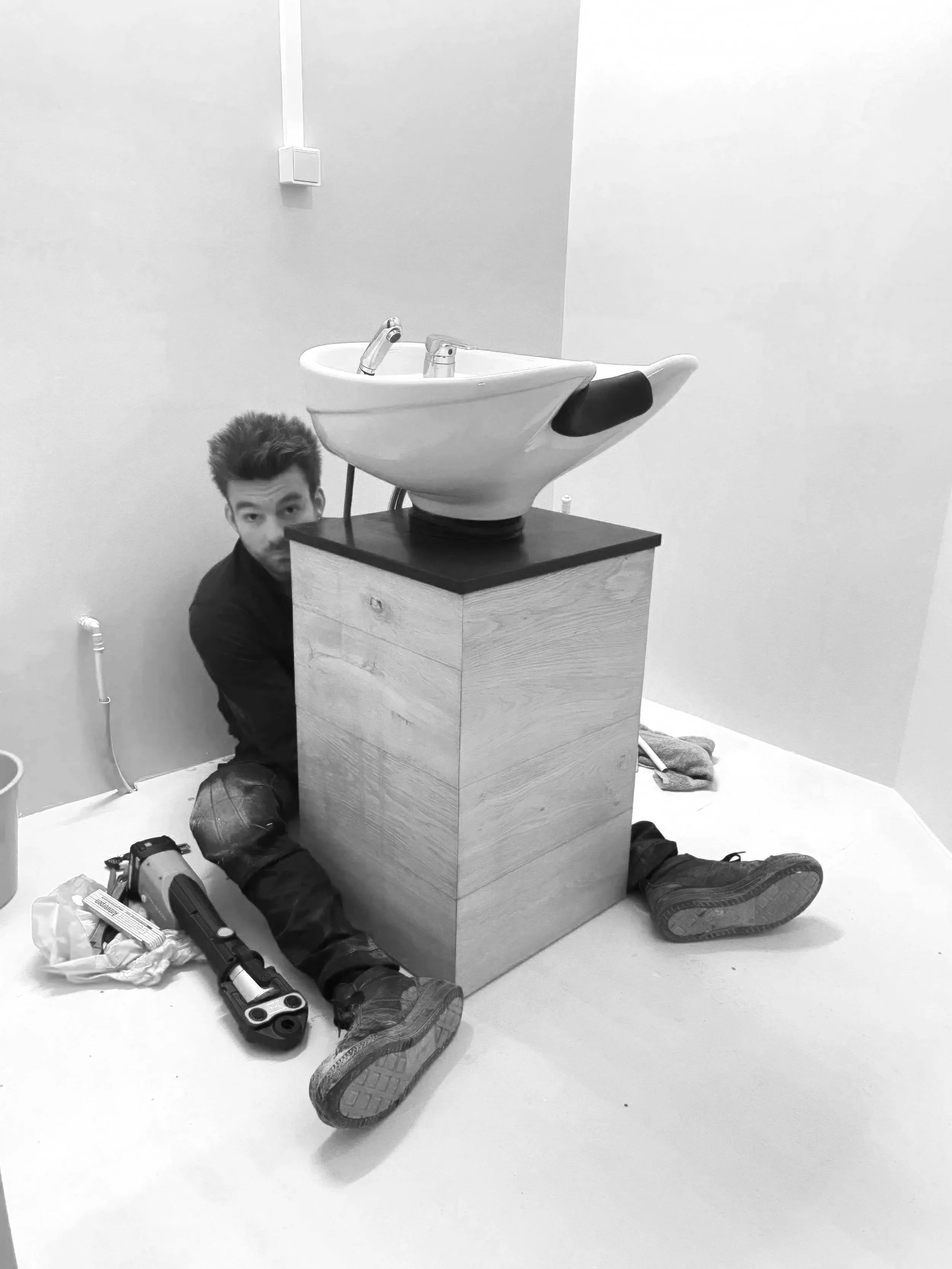 Een man zittend op de vloer naast een houten blok met daarop een wasbak die eruitziet als een paar schoenen.