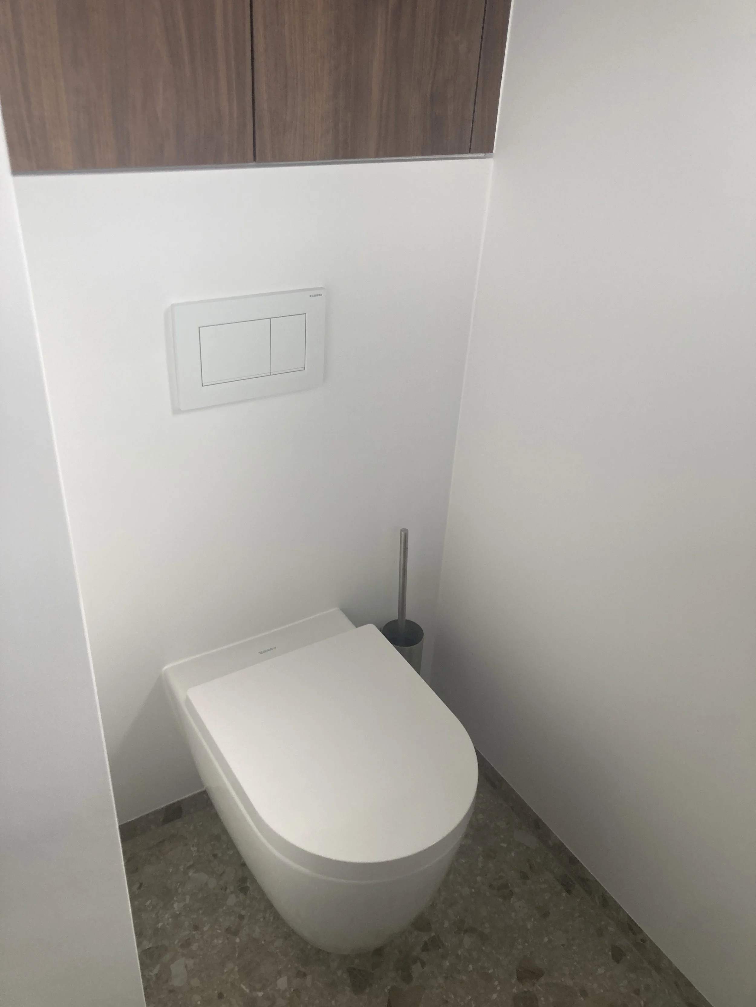 Een moderne wc met een wit toilet en een toiletborstel ernaast, ingebouwd in een minimalistische badkamer met witte muren en een houten kast.