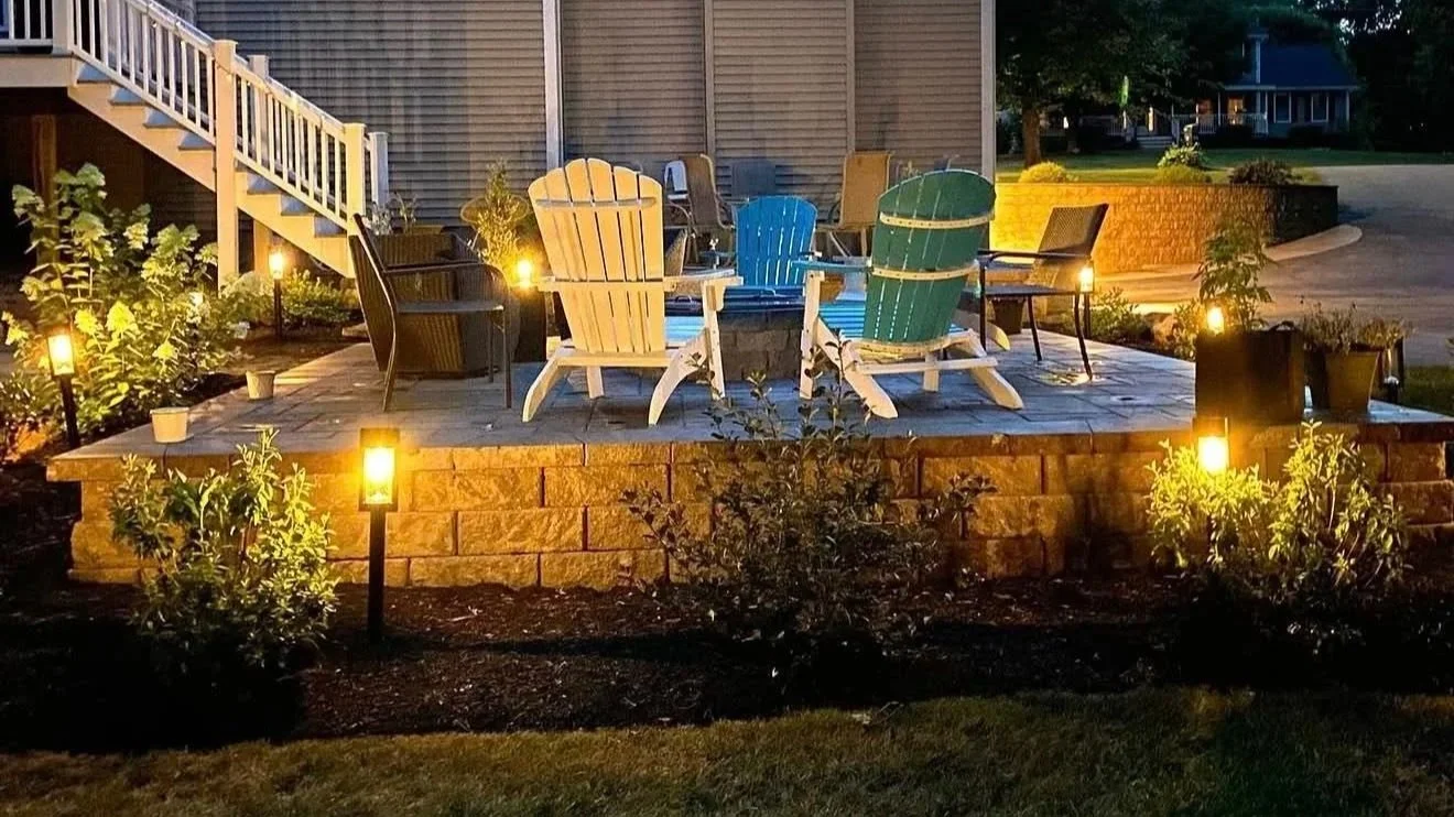 Nighttime outdoor patio with string lights, Adirondack chairs, and a small fire pit, surrounded by plants and landscape lighting.