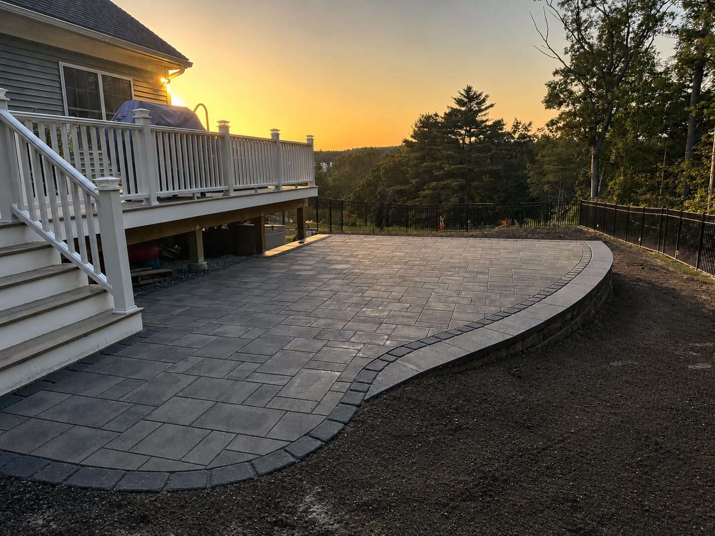 Backyard patio with newly installed gray paving stones, curved design, and black metal fence, adjacent to a house with a deck and stairs at sunset with trees in the background.