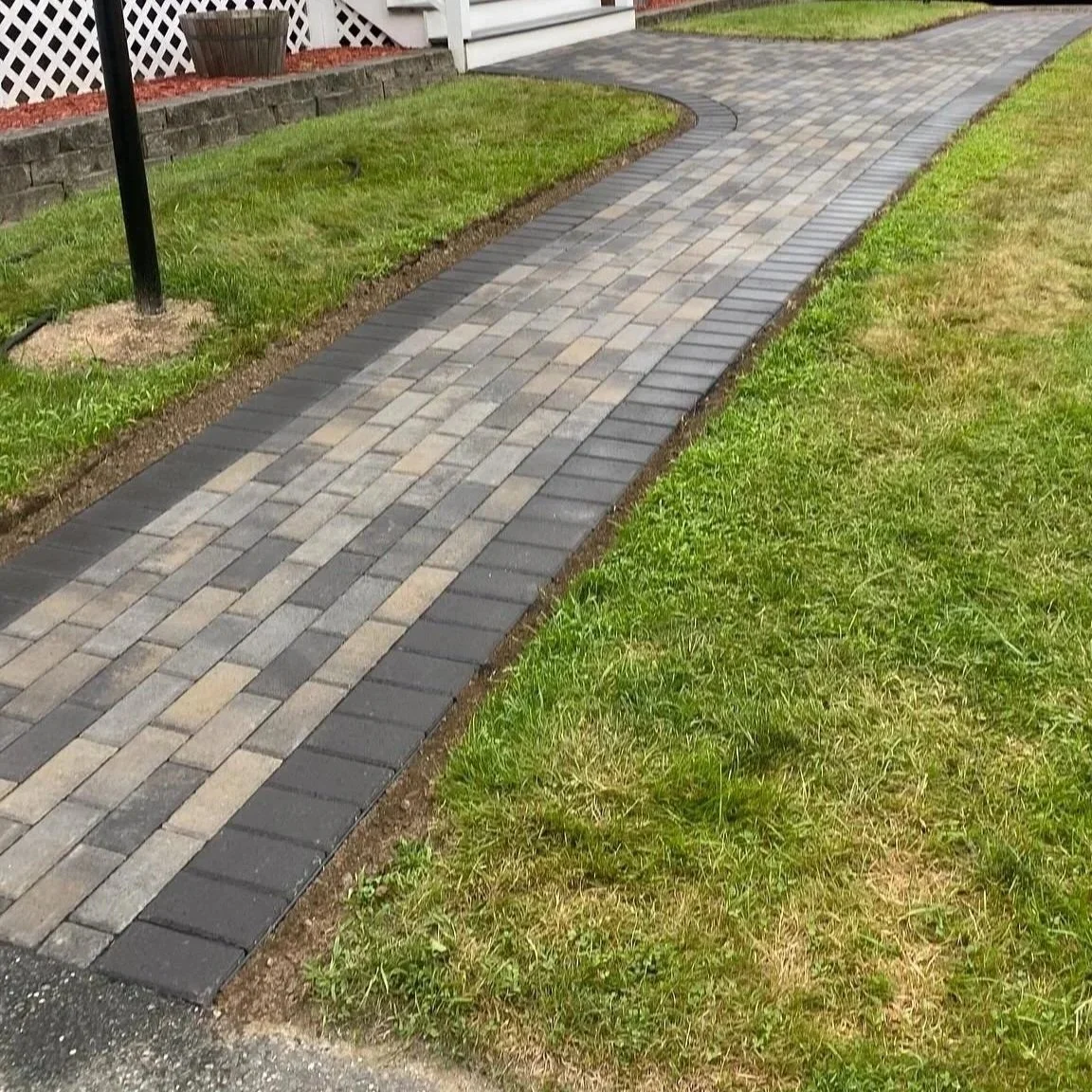 A curved paved walkway with gray, beige, and dark gray bricks, bordered by well-maintained grass on both sides, leading up to a house with a brick foundation and a white lattice structure.