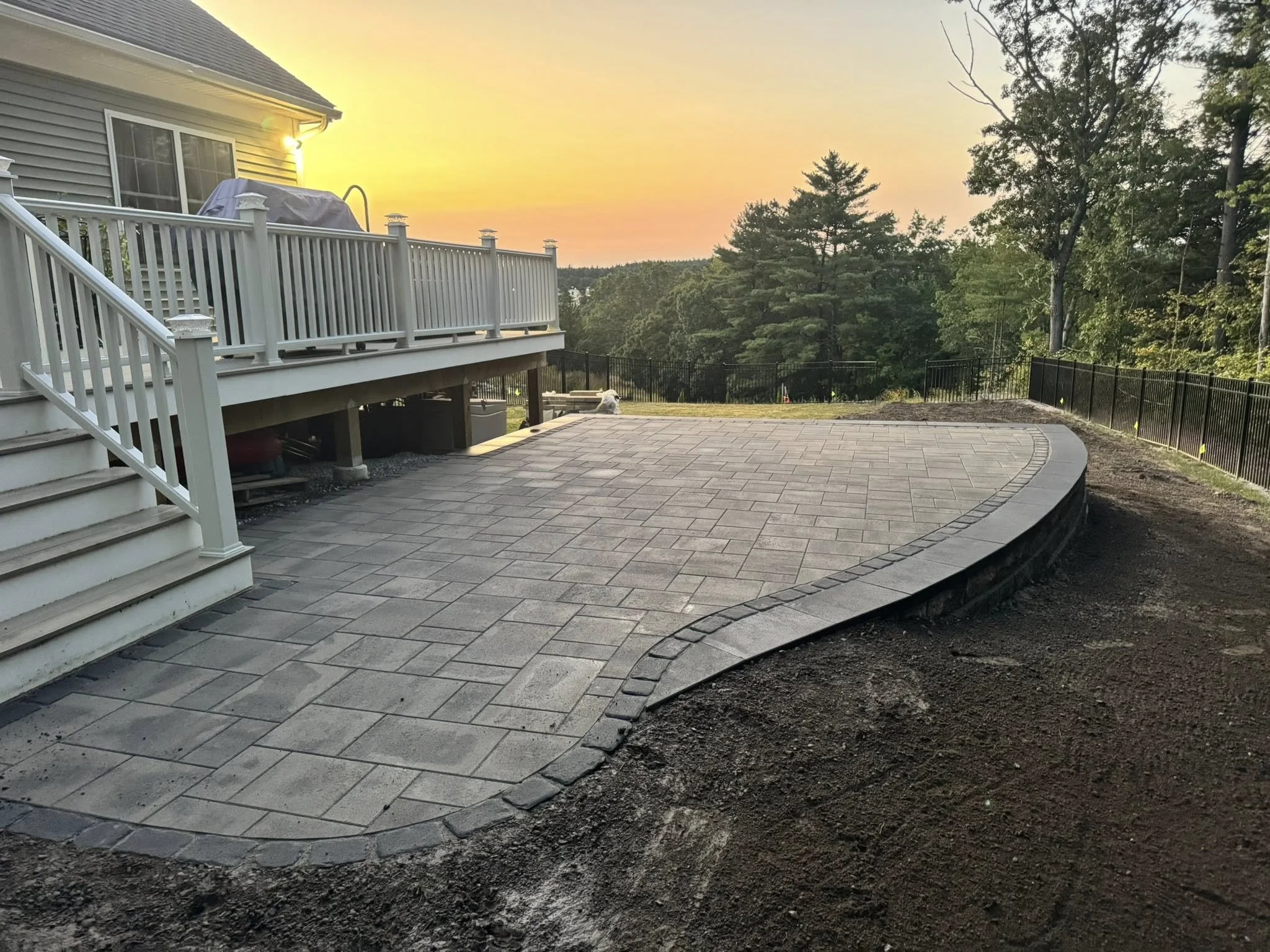 Newly paved patio with stone tiles, curved edges, and a white deck with stairs leading down to the patio. Black metal fence surrounds the yard, with trees and sunset in the background.