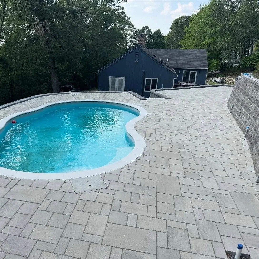 Newly finished backyard with a heart-shaped swimming pool, light-colored tiled deck, and a blue house with large windows surrounded by trees.