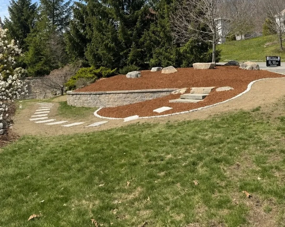 Landscaped yard with a curving pathway of white stones, a small set of stone steps, a retaining wall, mulch, and trees and shrubs in the background.