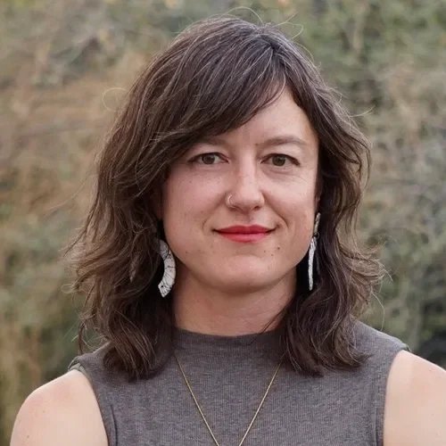 A woman with shoulder-length brown hair, wearing earrings, a gray sleeveless top, and a gold necklace, standing outdoors with trees in the background.