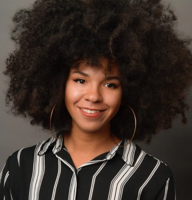 A woman with a big, curly Afro hairstyle, wearing large hoop earrings and a black and white striped shirt, smiling at the camera against a plain gray background.