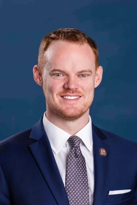 A smiling man in a navy suit, white shirt, and patterned tie, posing against a blue background.