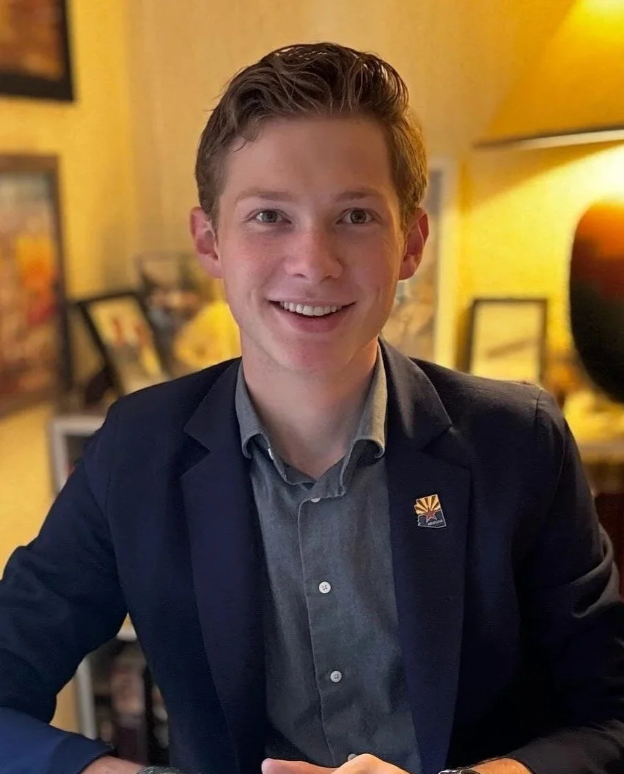Young man with light skin, light brown hair, smiling, wearing a dark blazer and a gray button-up shirt, sitting at a table in a warmly lit room with framed pictures and a lamp in the background.