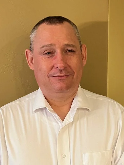 A man with short hair, wearing a white collared shirt, standing against a yellow wall.