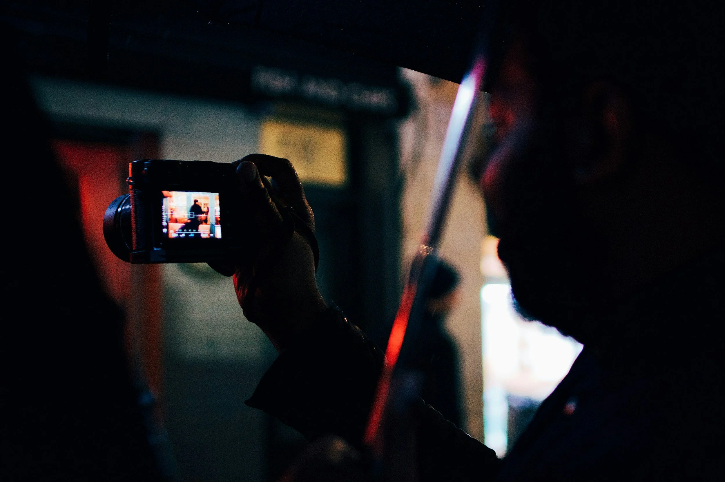 A person with dark hair and beard taking a photo in a dimly lit setting with a camera, with a blurred background including an illuminated sign or window.