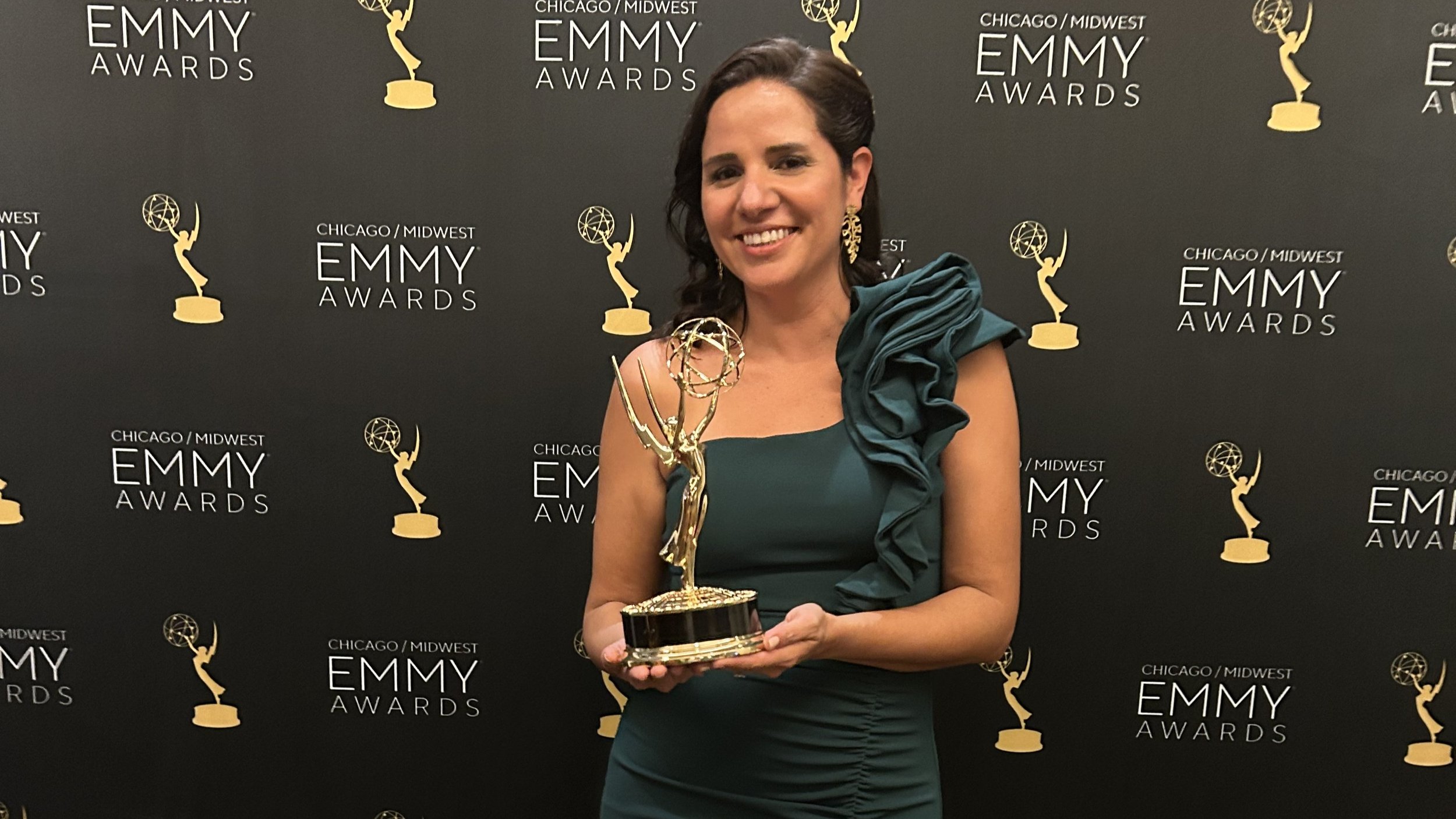 A woman in a dark green dress with ruffle shoulder detail, smiling and holding an Emmy Award trophy, standing in front of a black backdrop with Gold Emmy statues and the text "Chicago / Midwest Emmy Awards."