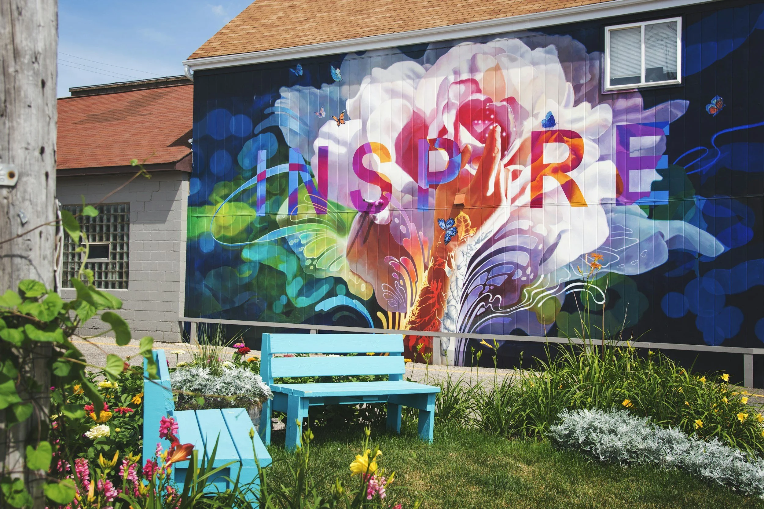 Colorful mural on a building wall with the word 'INSPIRE', a hand reaching towards a large pink flower, surrounded by butterflies and swirling patterns, with a blue bench and garden with flowers in the foreground.