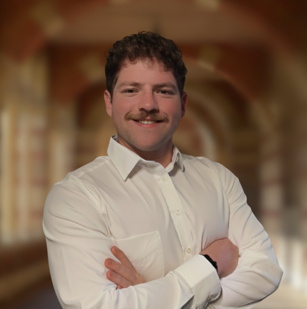 A man with brown hair, a mustache, and a white dress shirt smiling with arms crossed, standing in front of a blurred architectural background.