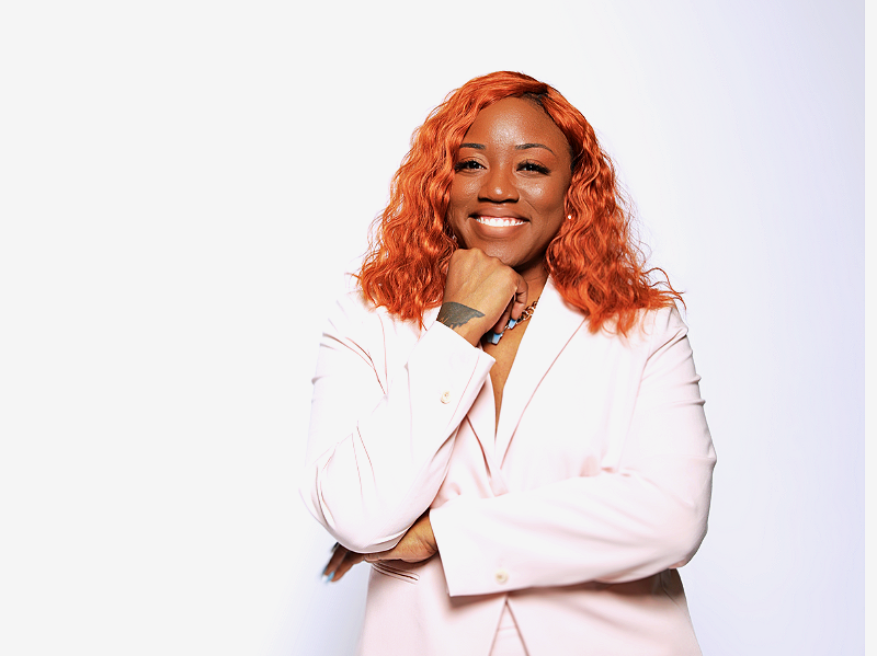 A smiling woman with curly orange hair wearing a light pink blazer, standing against a plain white background.