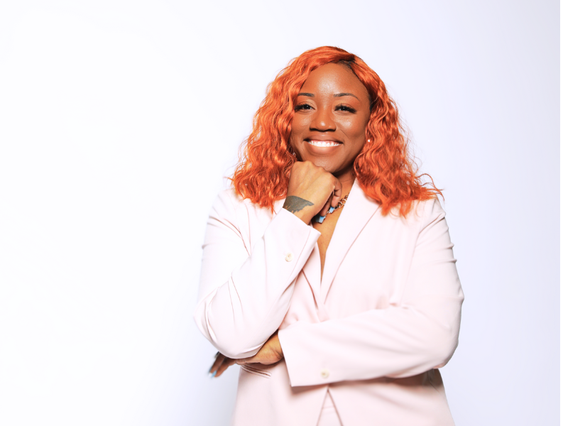 Portrait of a woman with curly orange hair smiling with her arms crossed and resting her chin on her hand, wearing a light pink blazer against a white background.