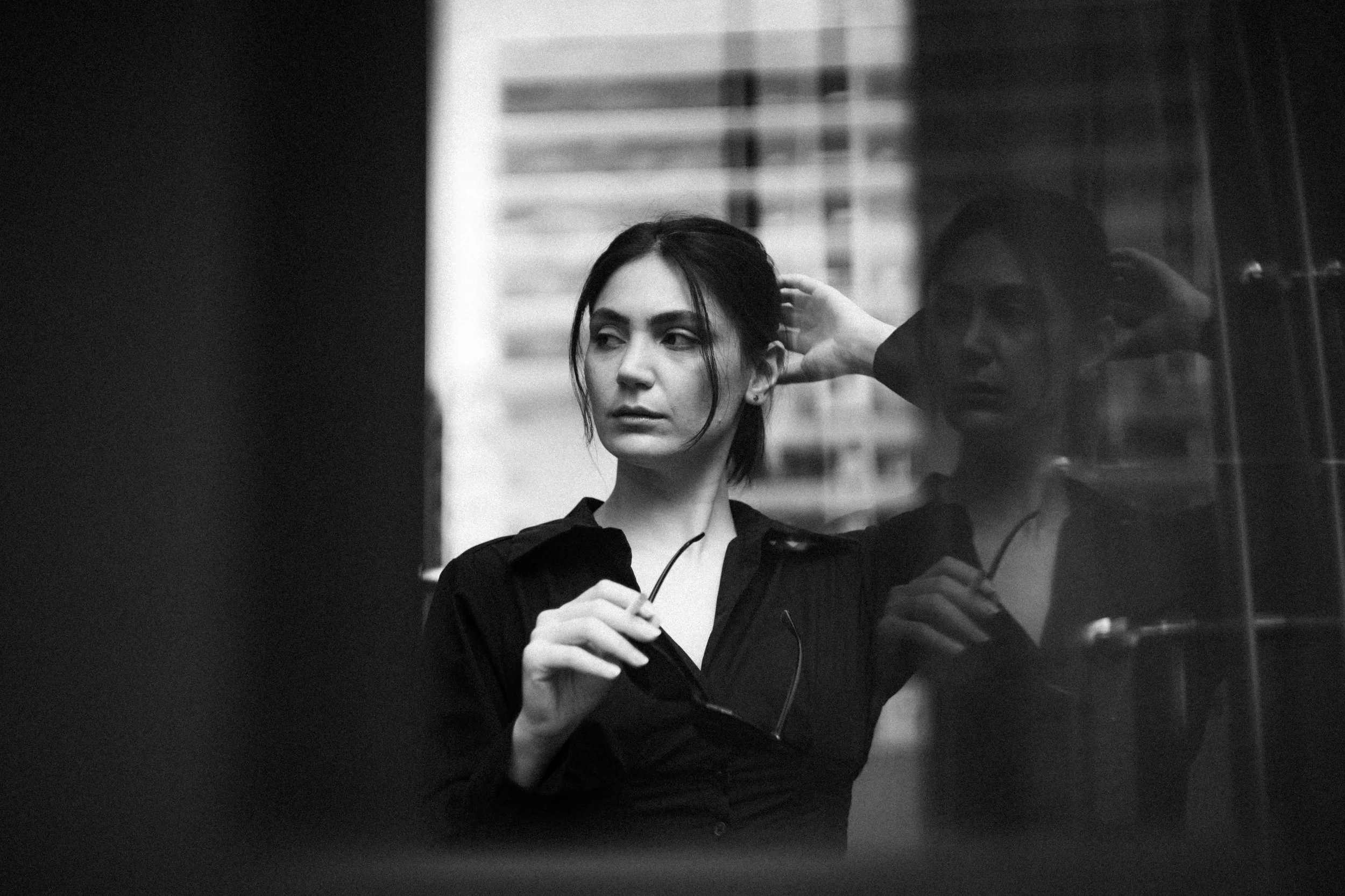 A black and white photo of a woman looking out a window, holding her glasses, with a cityscape reflected behind her.