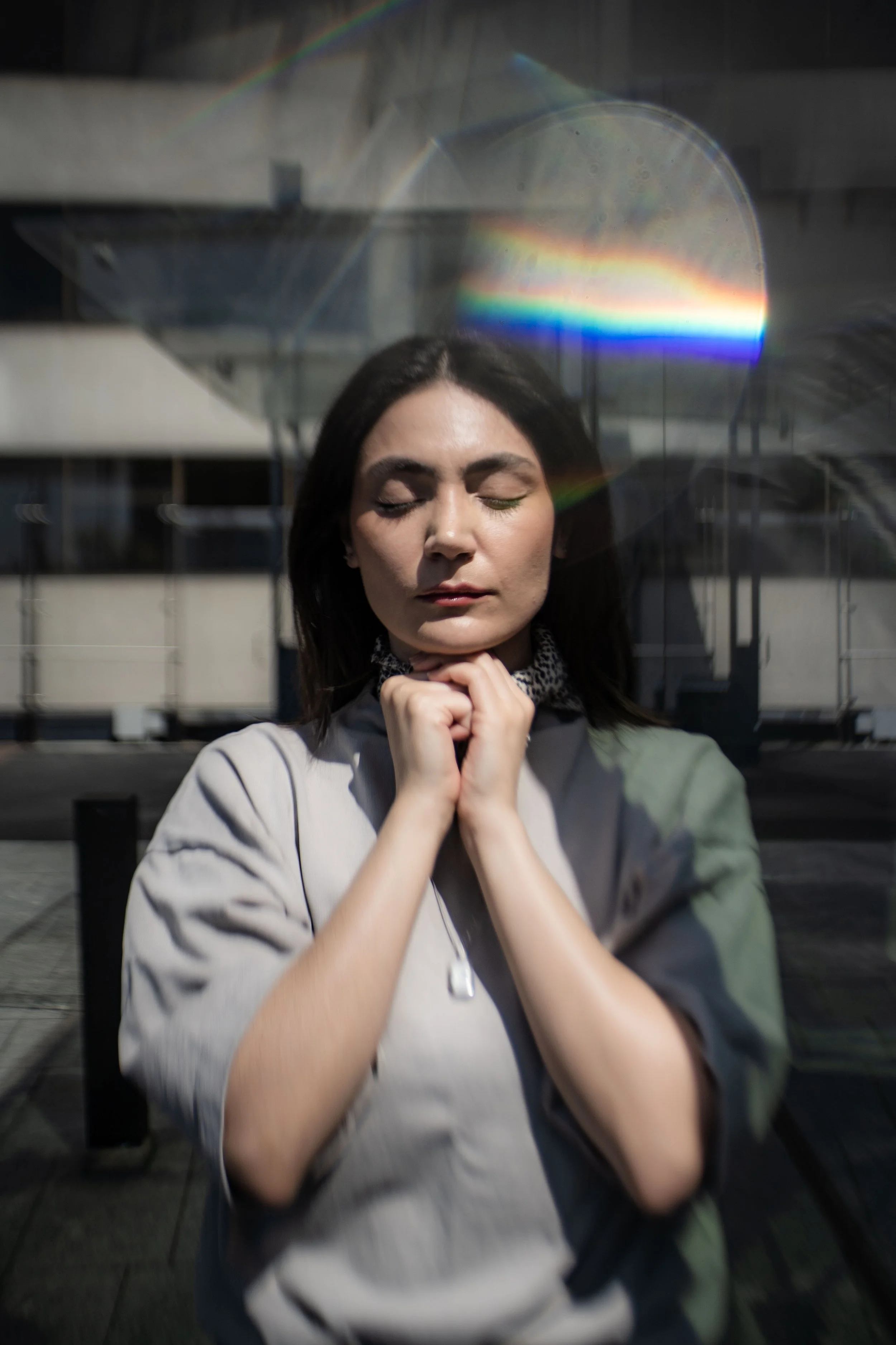 A woman with dark hair and closed eyes standing outdoors, with sunlight creating a rainbow reflection on a glass surface in front of her.