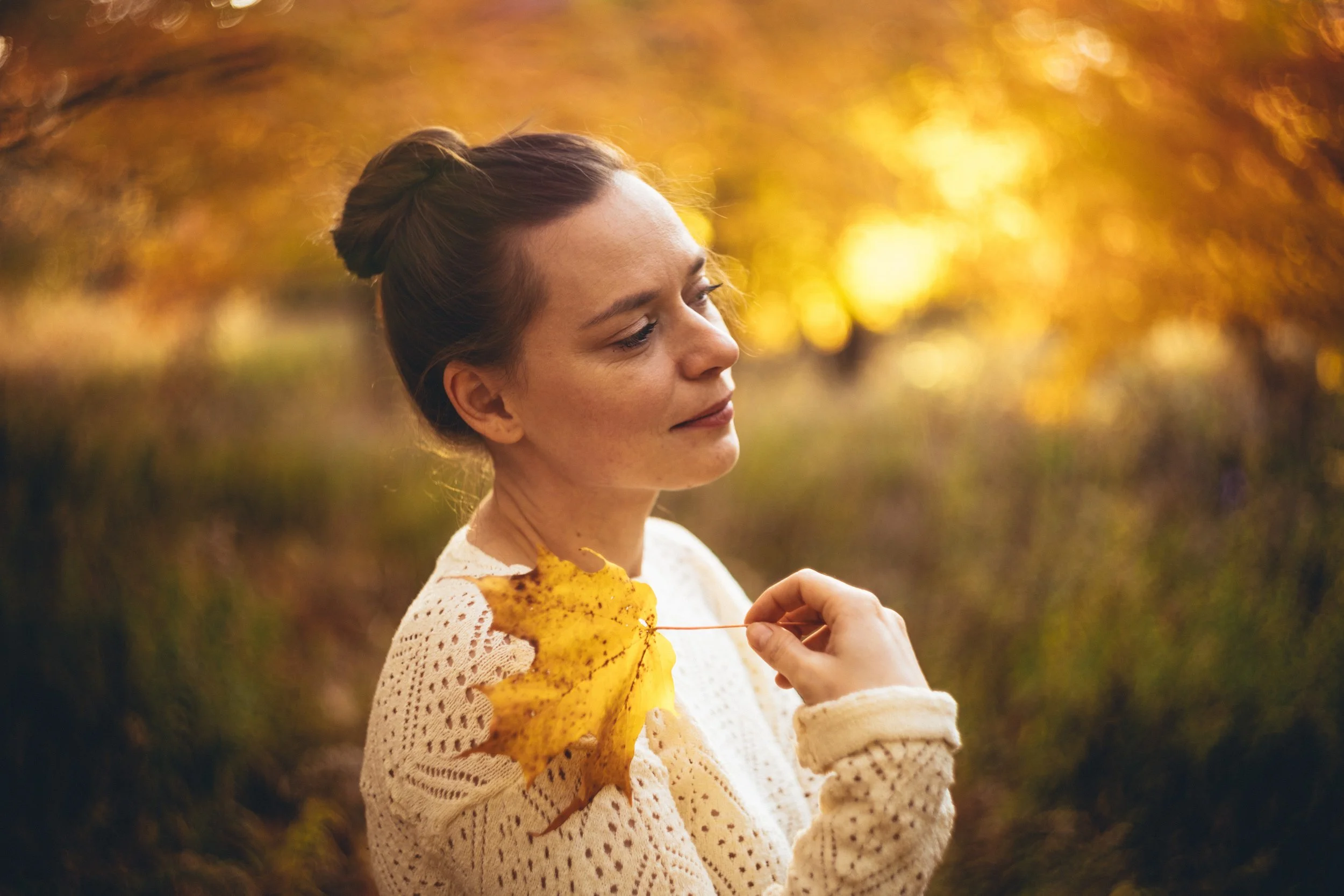 A woman with brown hair tied in a bun, wearing a cream-colored knit sweater, stands outdoors during autumn. Someone is gently pinning a large yellow autumn leaf to her shoulder. The background is filled with blurred yellow and orange autumn trees, il