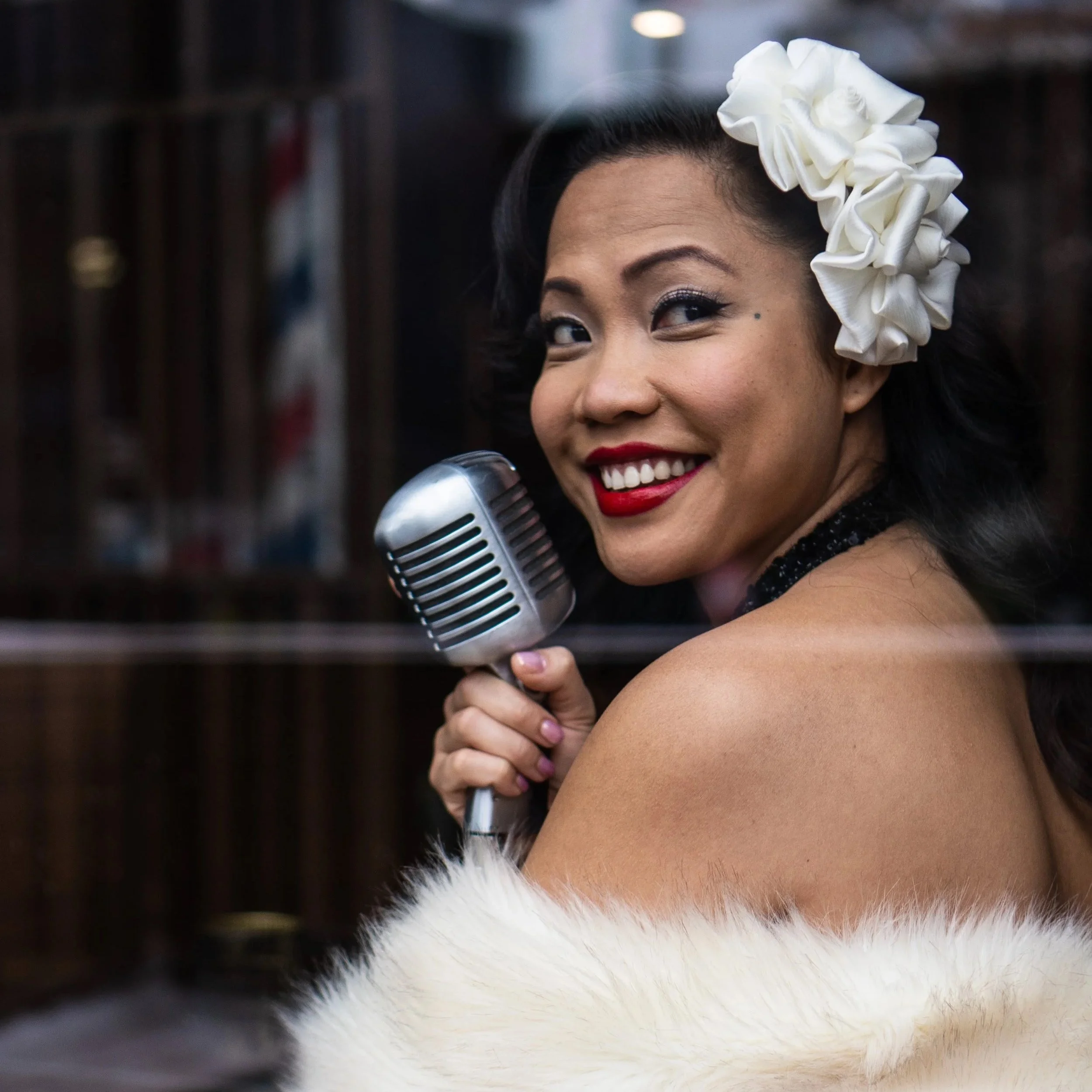 A woman with vintage style makeup and a white floral hair accessory, smiling while holding a microphone.