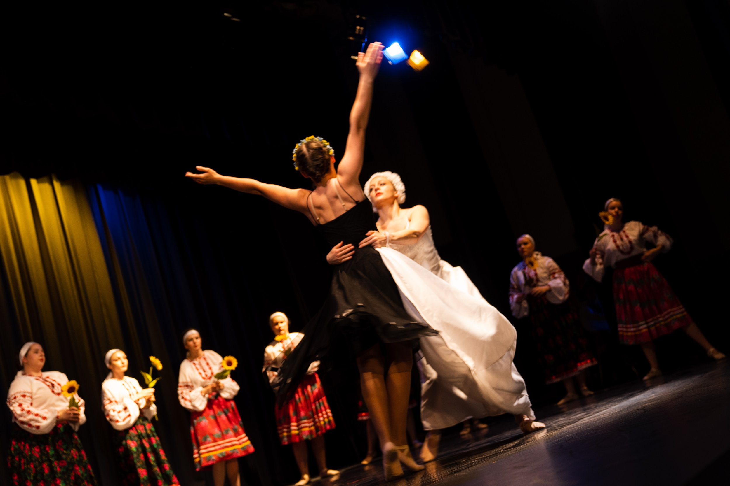 Dancers in traditional folk costumes performing on stage, with one dancer in a black dress and another in a white dress closely engaged, surrounded by other dancers in embroidered blouses and colorful skirts.