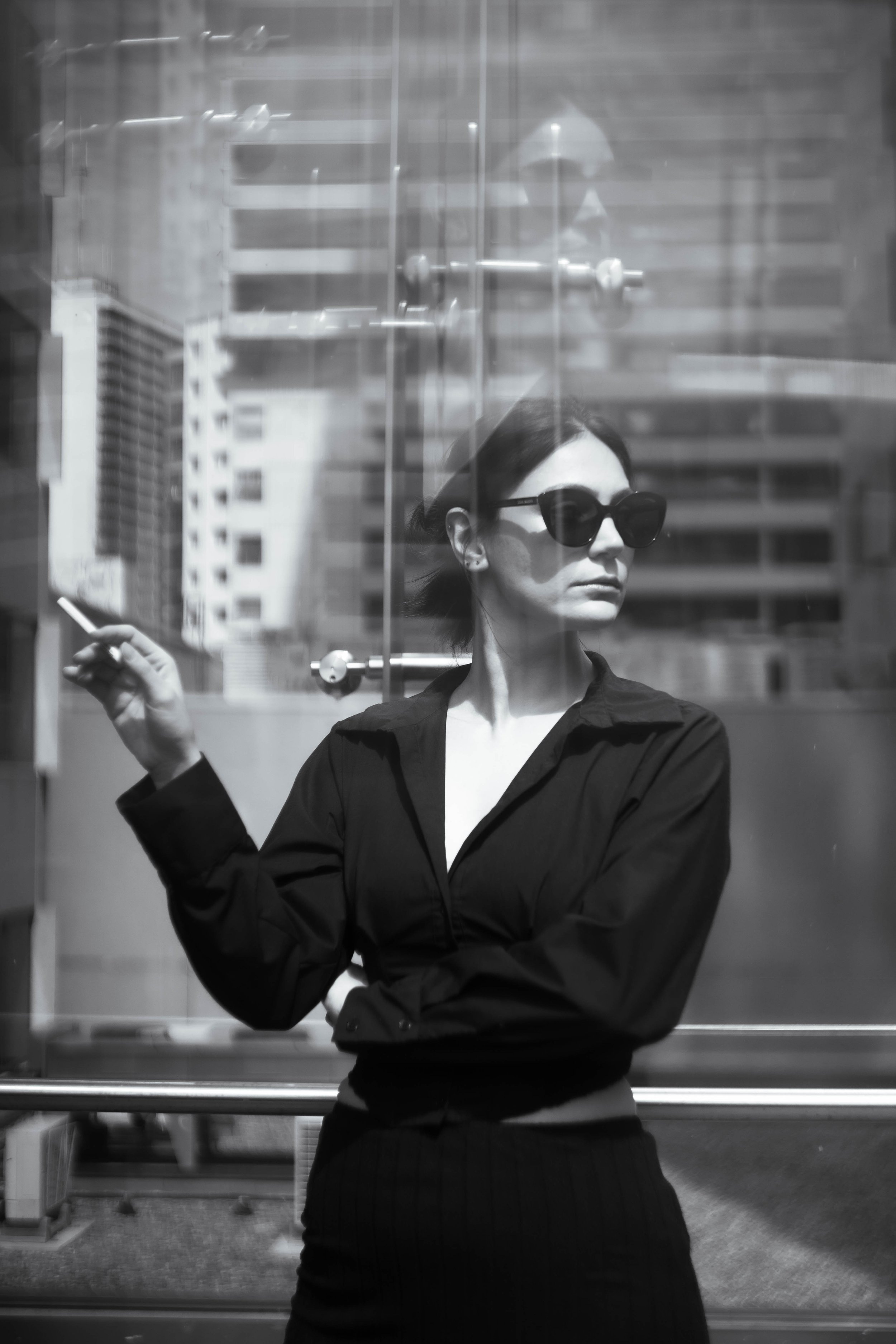 A woman wearing sunglasses and a black shirt stands behind a glass window, holding a cigarette in her right hand with a city skyline in the background.