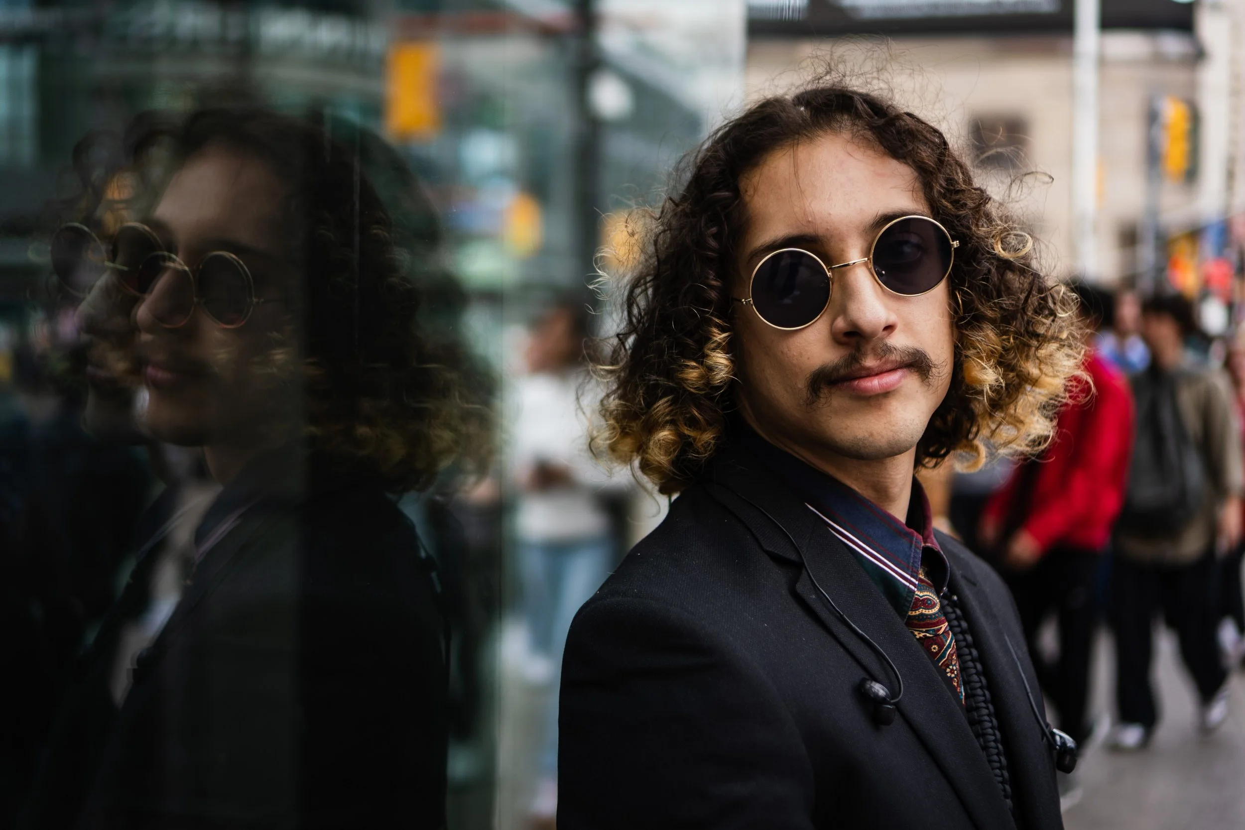 A man with curly hair and round sunglasses standing next to a reflective glass wall in a city street with blurred pedestrians in the background.