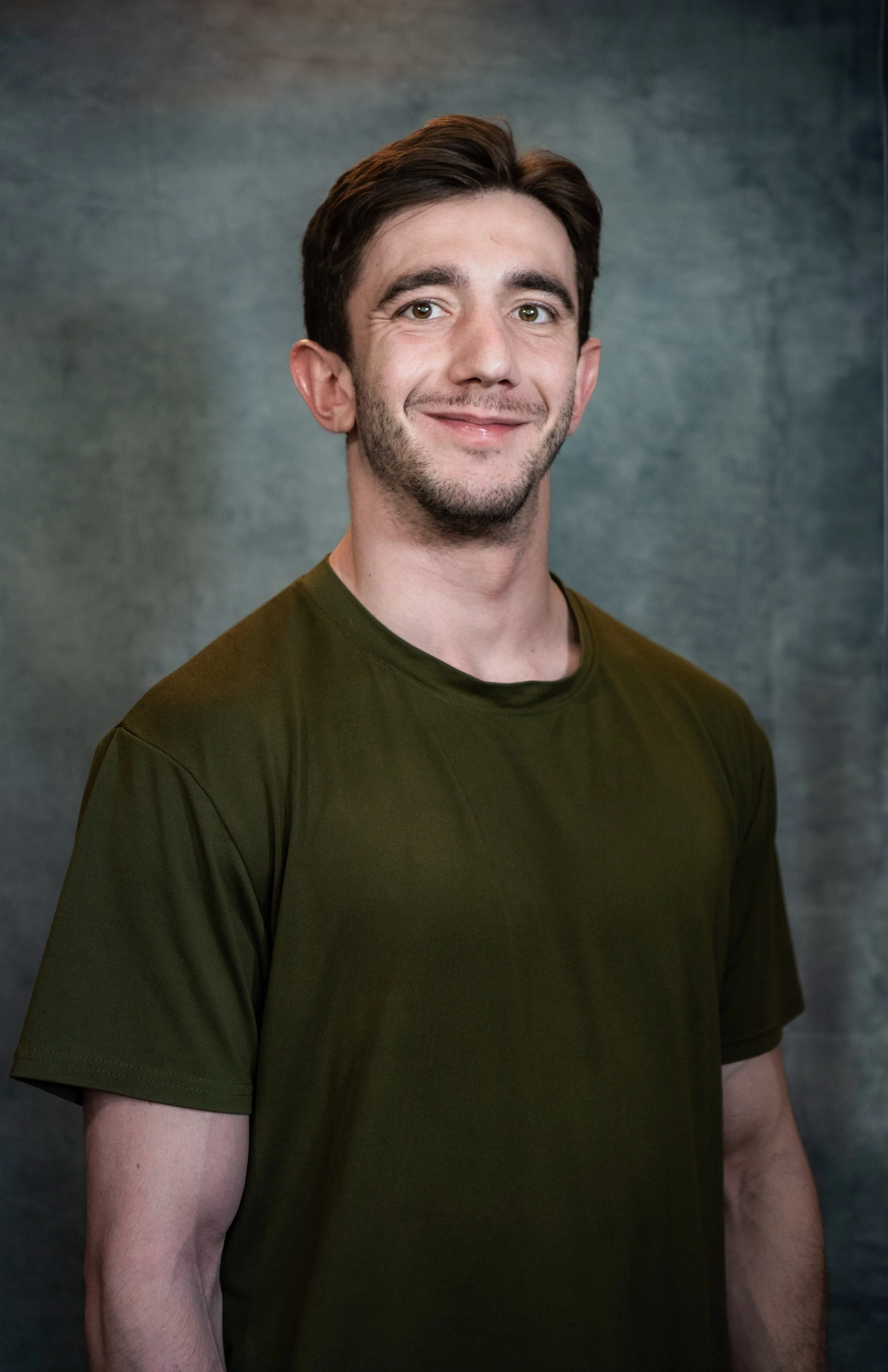 A young man with dark hair, facial hair, and a slight smile, wearing a dark green t-shirt, standing against a gray background.