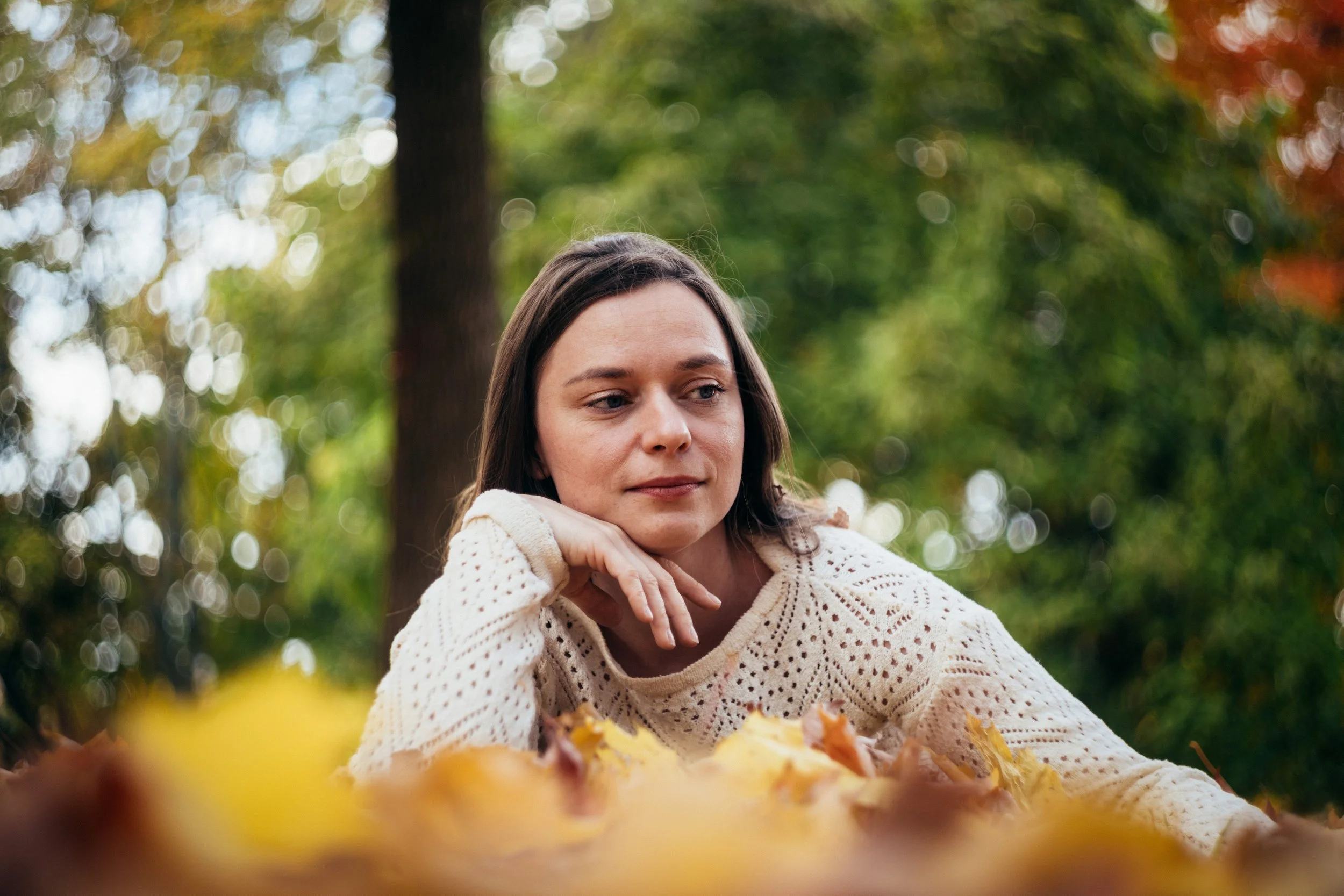 A woman with dark hair and a beige knit sweater resting her chin on her hand, lying among fallen autumn leaves in a park with trees and green autumn foliage in the background.