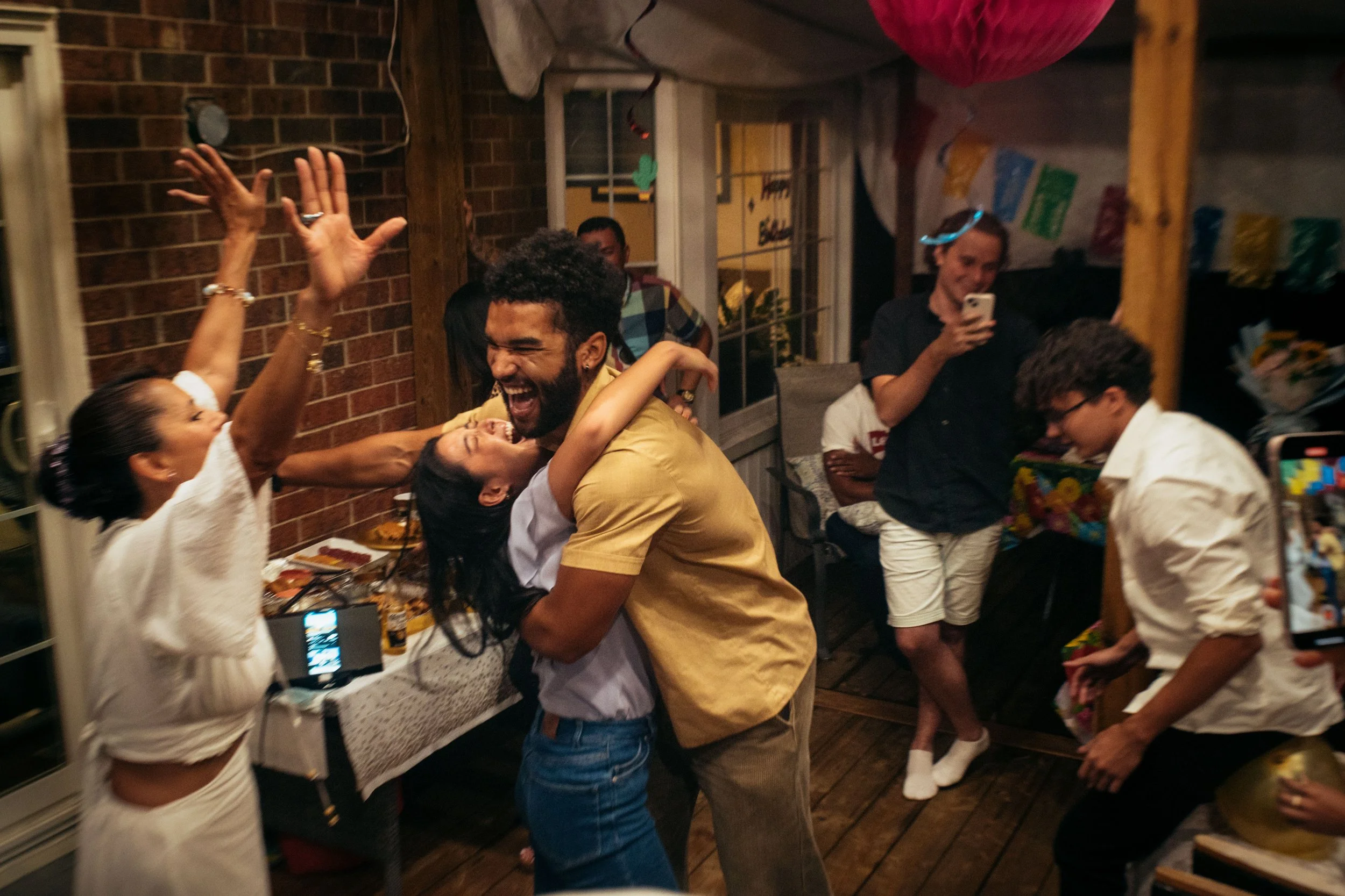 People celebrating at a party, dancing, hugging, and taking photos indoors with decorations.