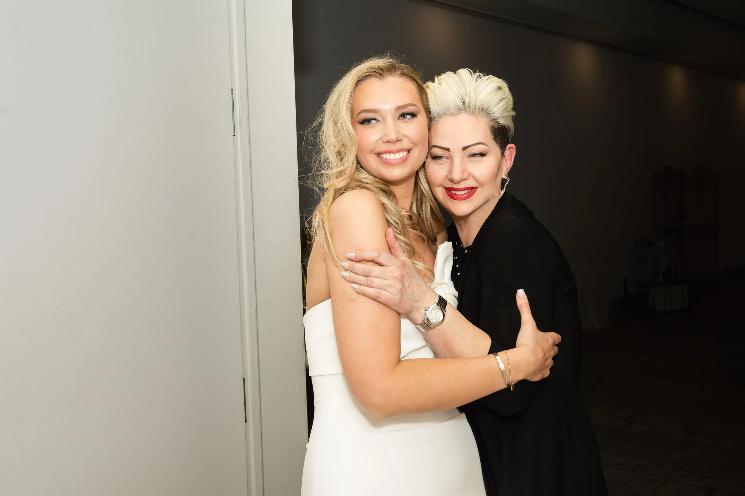 Two women hugging and smiling, one in a white dress and the other in black, indoors with a dark background.