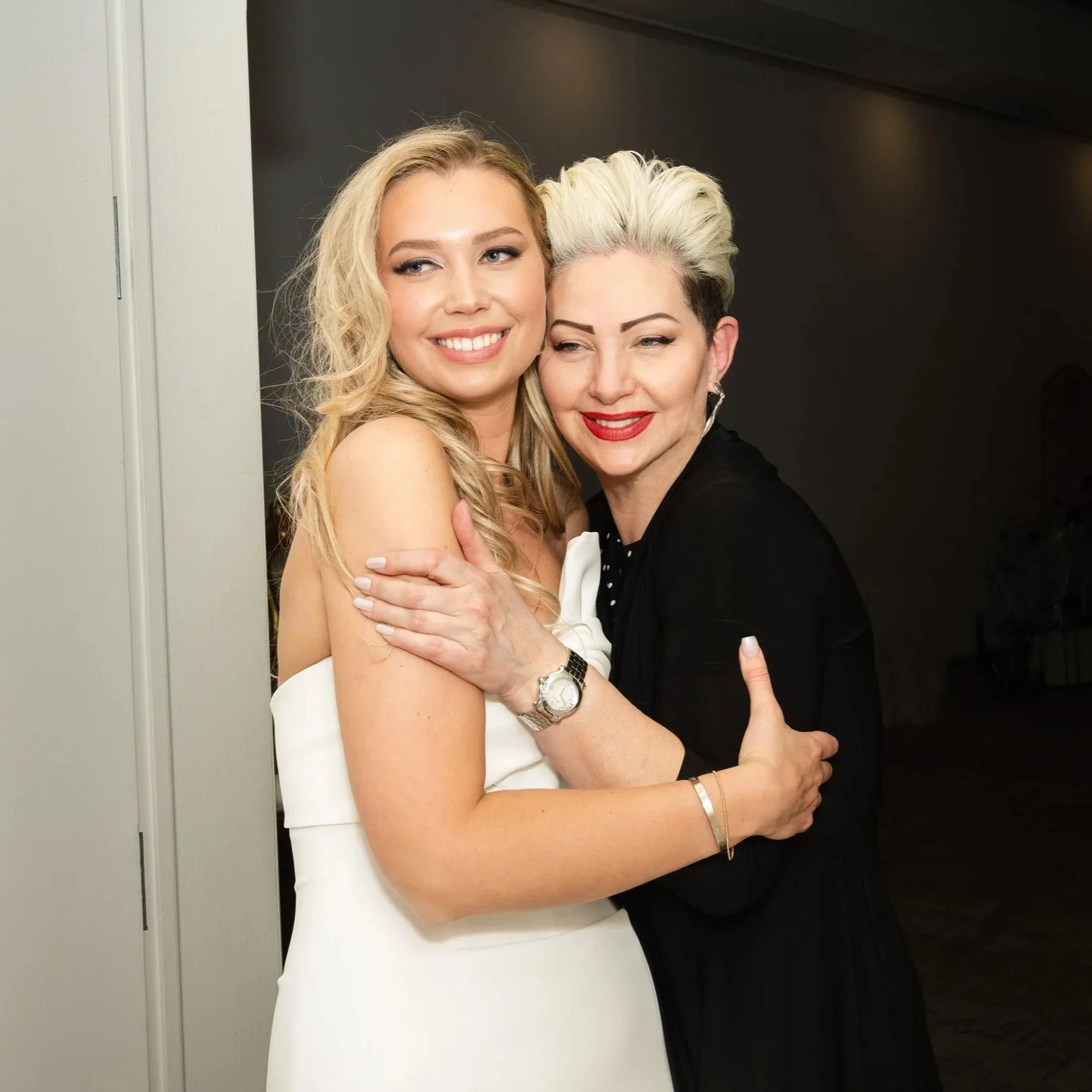 Two women hugging and smiling for the camera; one woman has blonde hair and the other has platinum blonde hair with dark roots.