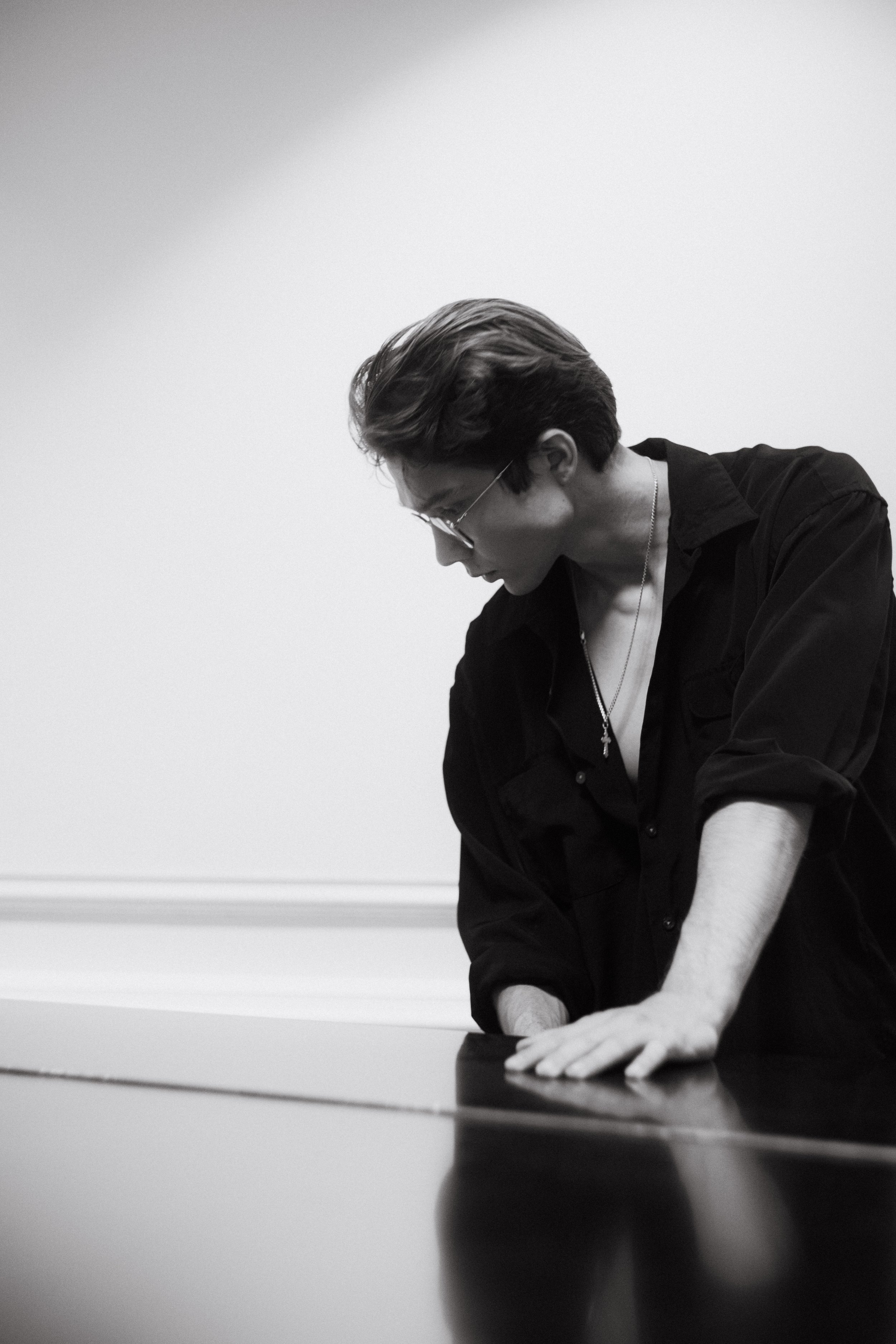 A black and white photograph of a person with short hair, glasses, and a necklace, leaning over and placing their hands on a flat surface, with a plain wall in the background.