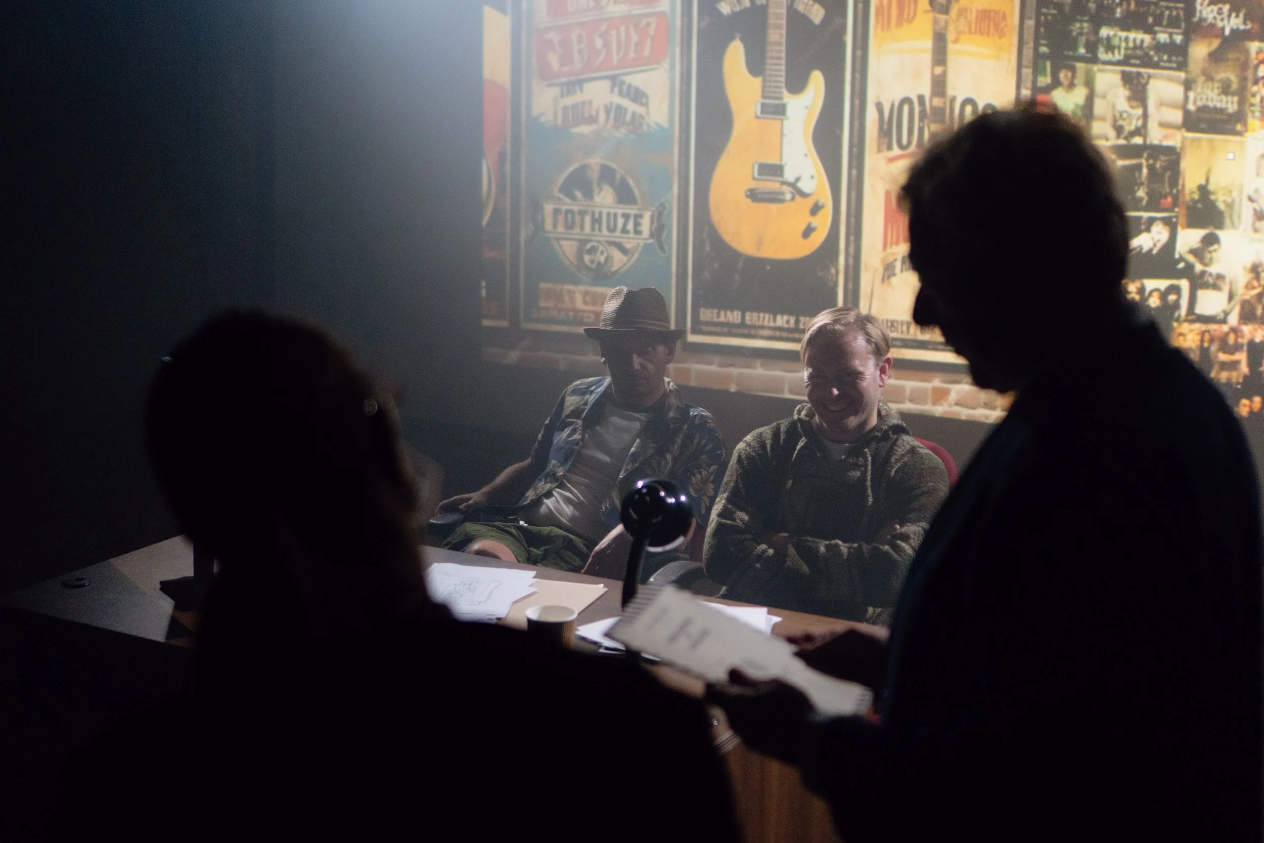 Four people in a dark room with a brick wall decorated with posters, two are sitting on the side and two are standing, with one of them reading from papers.