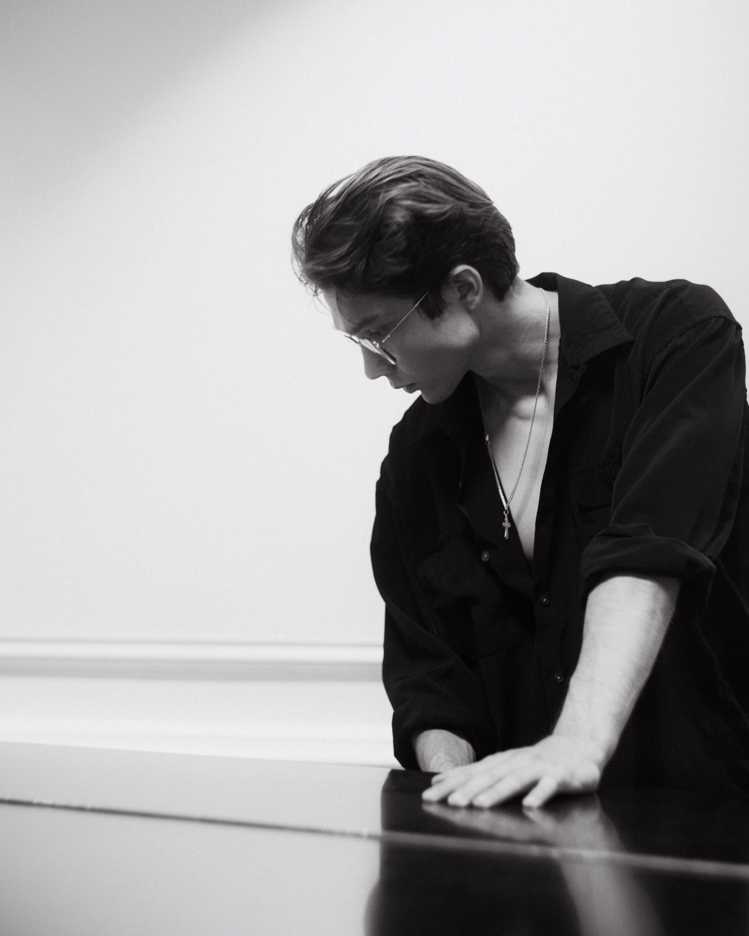 Black and white photo of a person with short hair, glasses, and a necklace, leaning over a table with their hand on the surface.