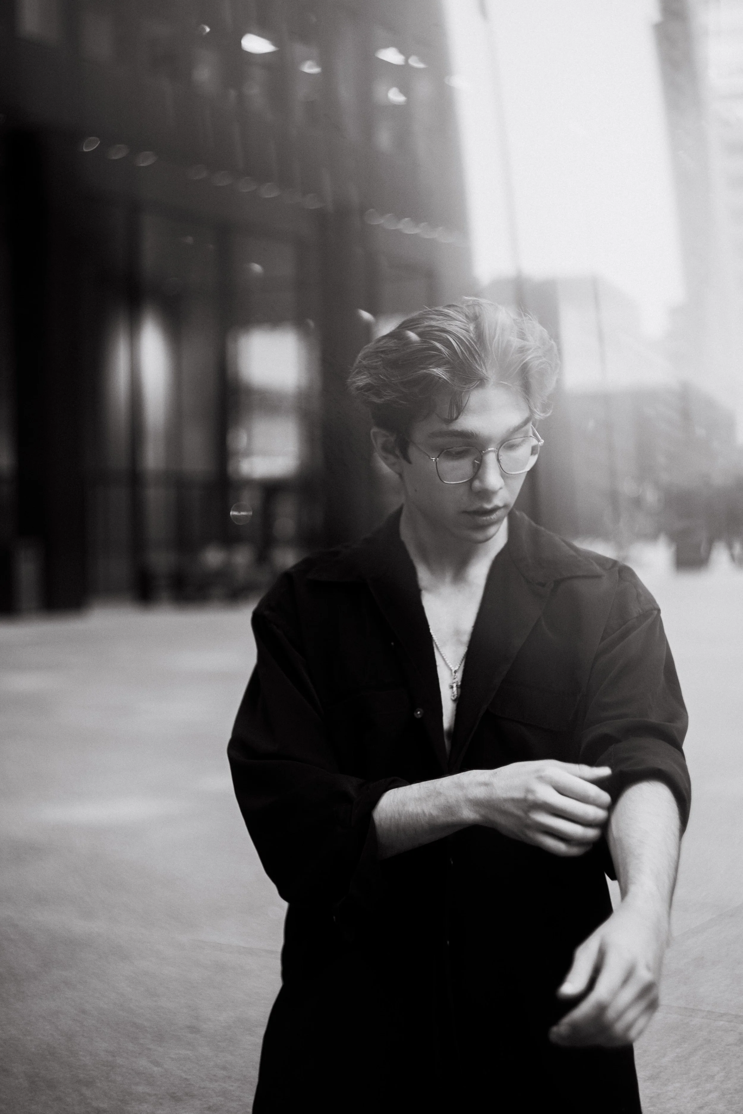 A young man with glasses and styled hair is outdoors, adjusting his rolled-up sleeve, with urban buildings and a reflective glass building in the background, captured in black and white.
