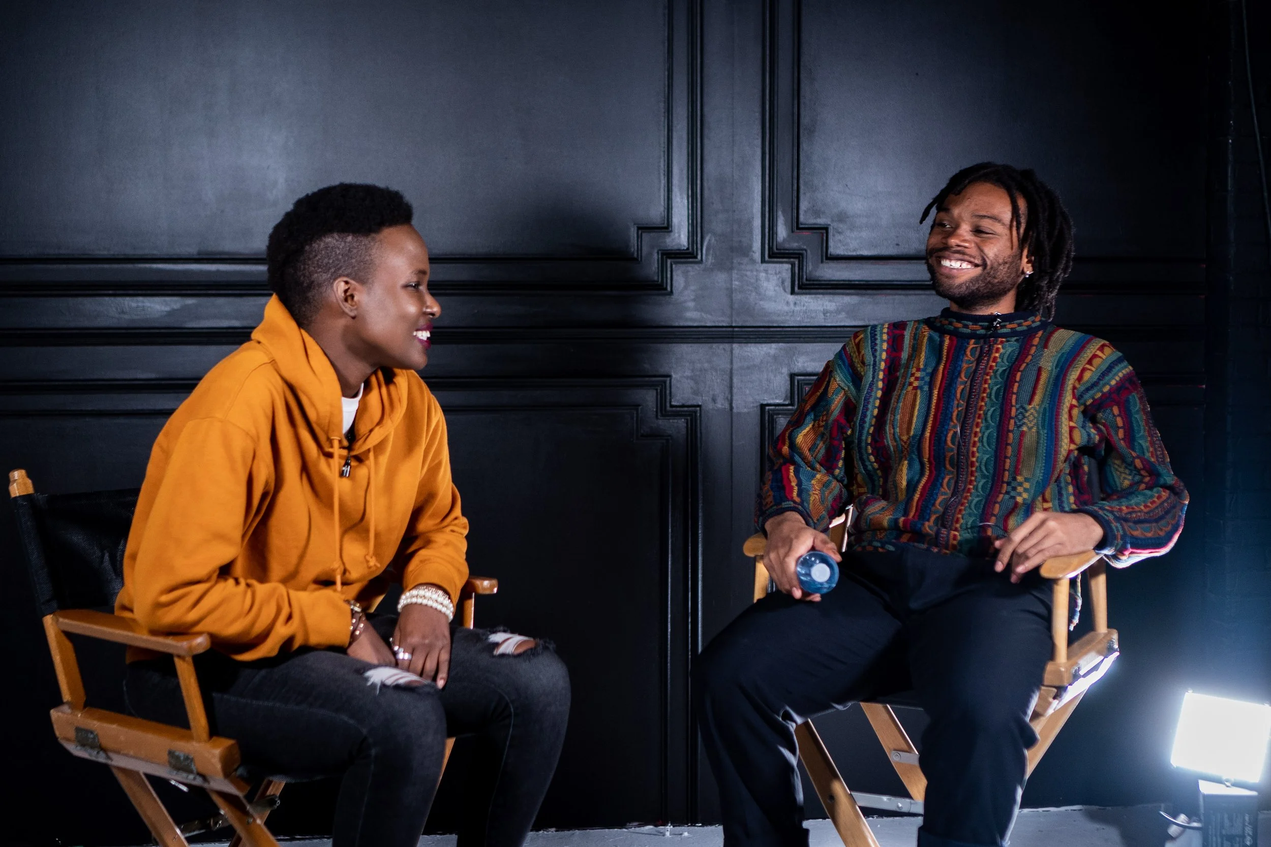Two people sitting on chairs, smiling and talking, with a black wall background and a bright light on the floor.