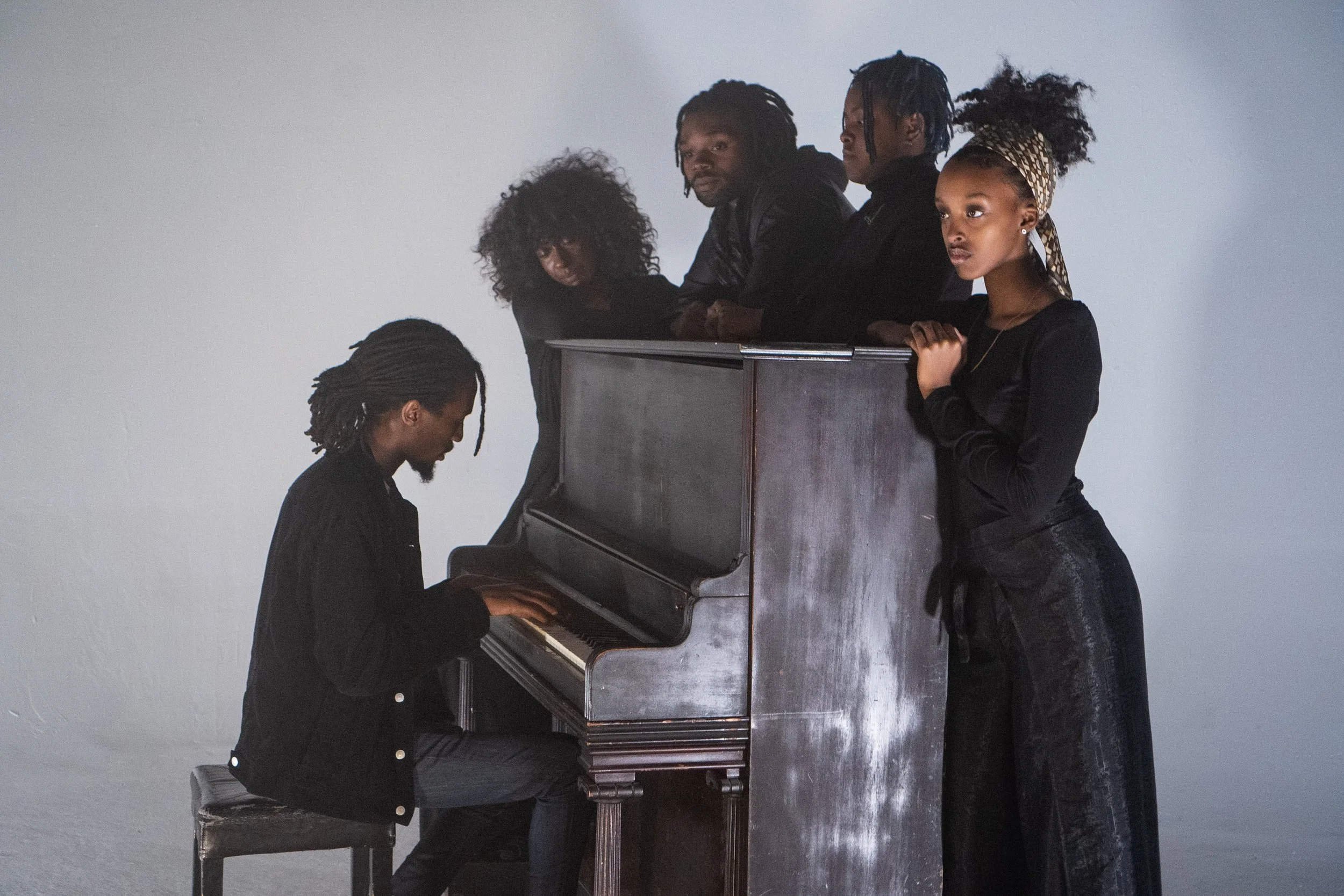 A group of six African American people, with one playing a grand piano and the others gathered around, looking on.