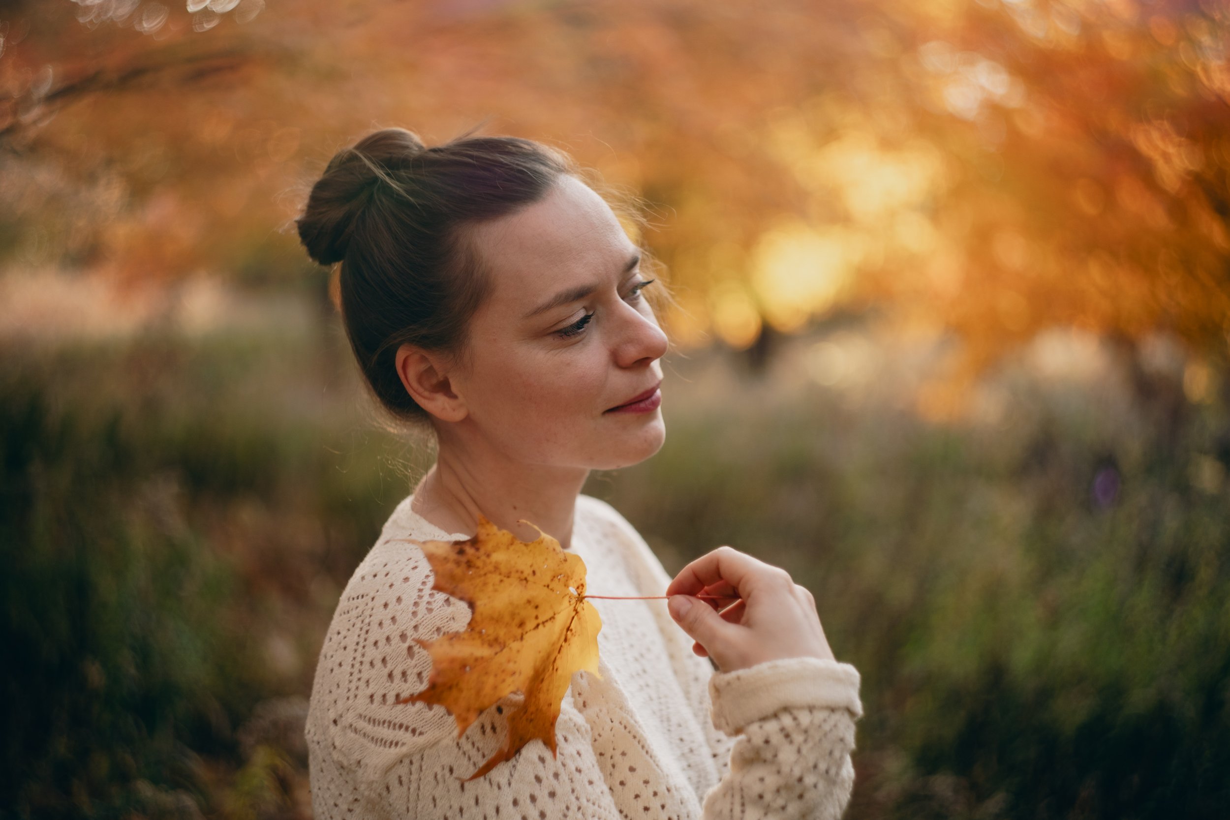 A woman holding a yellow autumn leaf with a background of orange fall foliage.