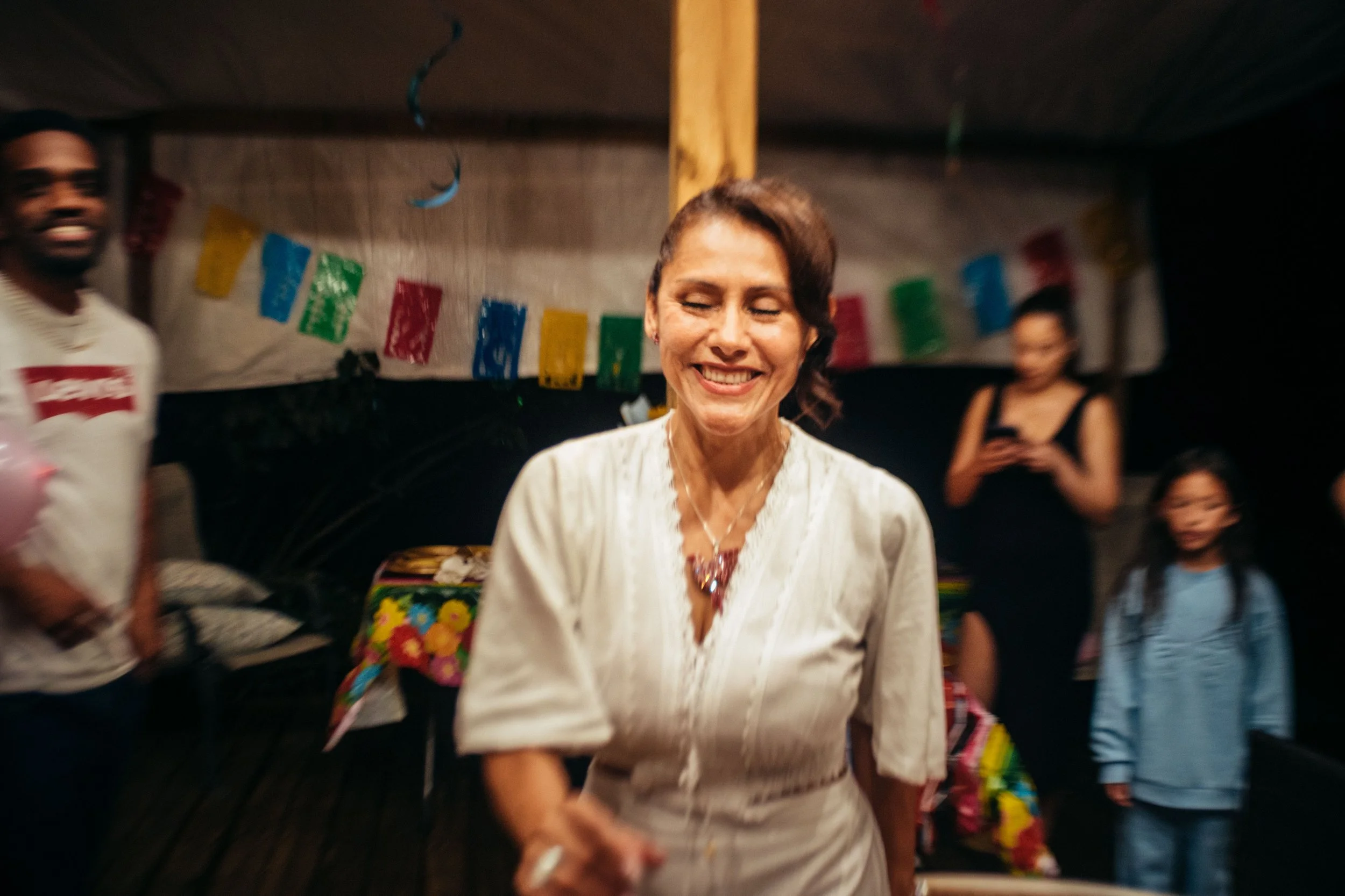 A woman with short brown hair, smiling with her eyes closed, wearing a white blouse and a necklace, at a festive gathering with colorful banners and other people in the background.