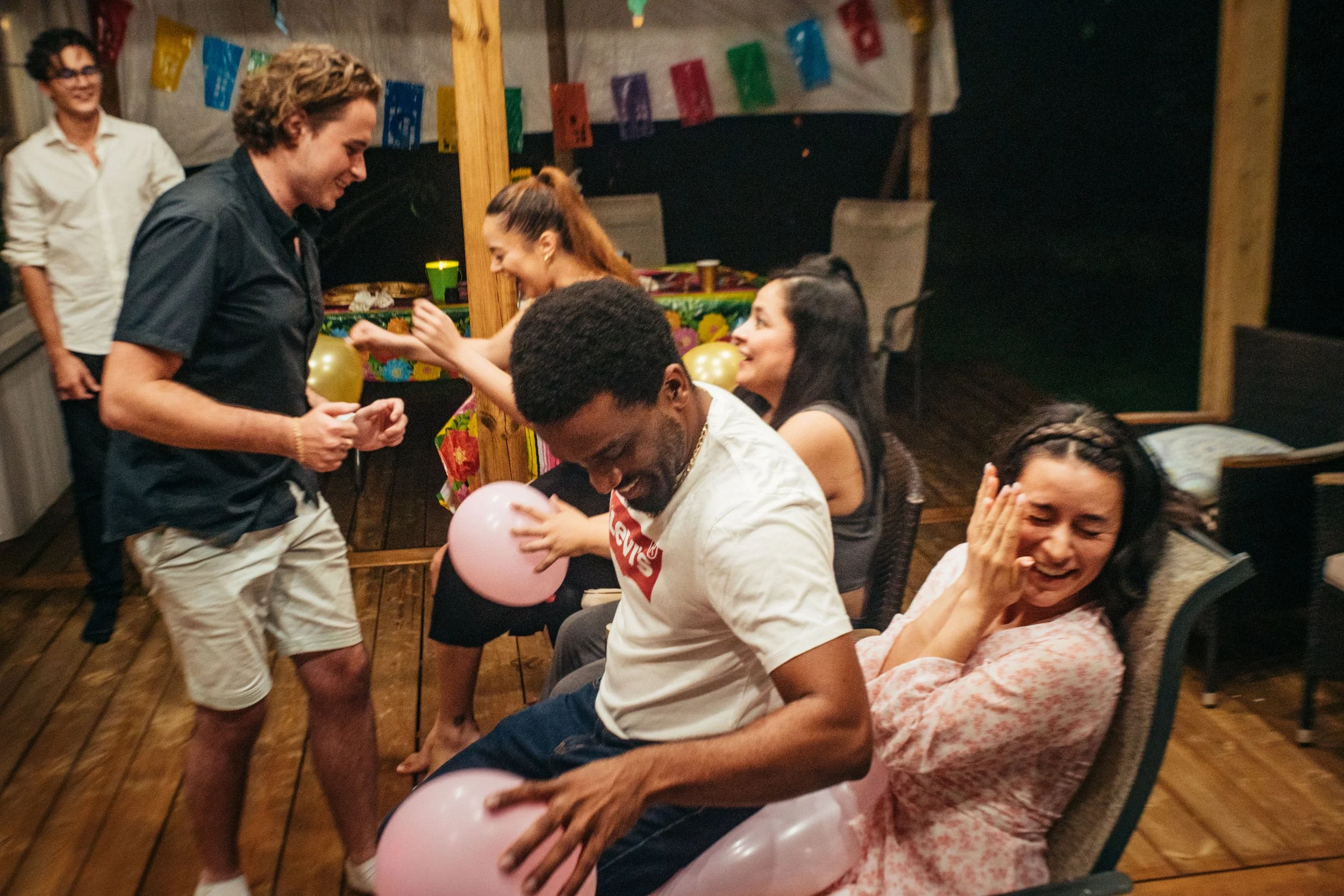 A group of people celebrating a birthday party indoors, with some sitting and some standing, holding balloons and laughing.