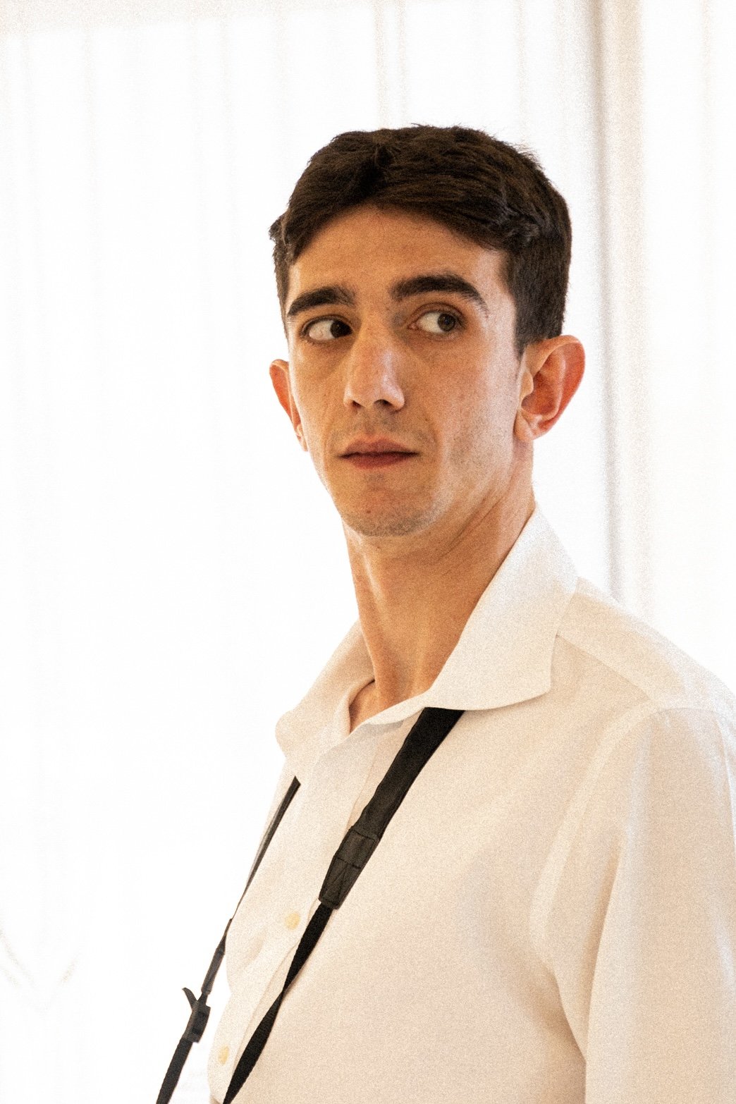 A young man with dark hair wearing a white shirt, looking to the side with a camera hanging around his neck, standing against a bright, light-colored background.