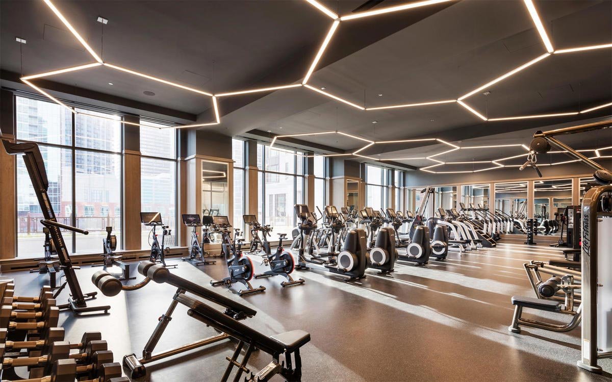 Modern gym interior with exercise bikes, treadmills, weight machines, and free weights, featuring geometric ceiling lights and floor-to-ceiling windows.
