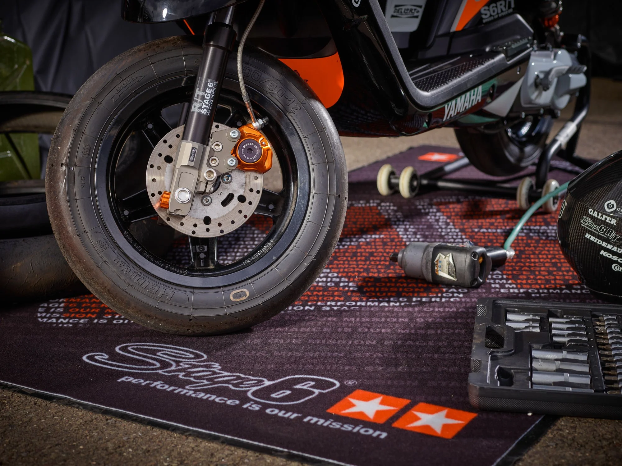 Close-up of a racing motorcycle front wheel, with a black tire, brake disc, and orange brake caliper, on a black mat with racing branding and tools nearby.