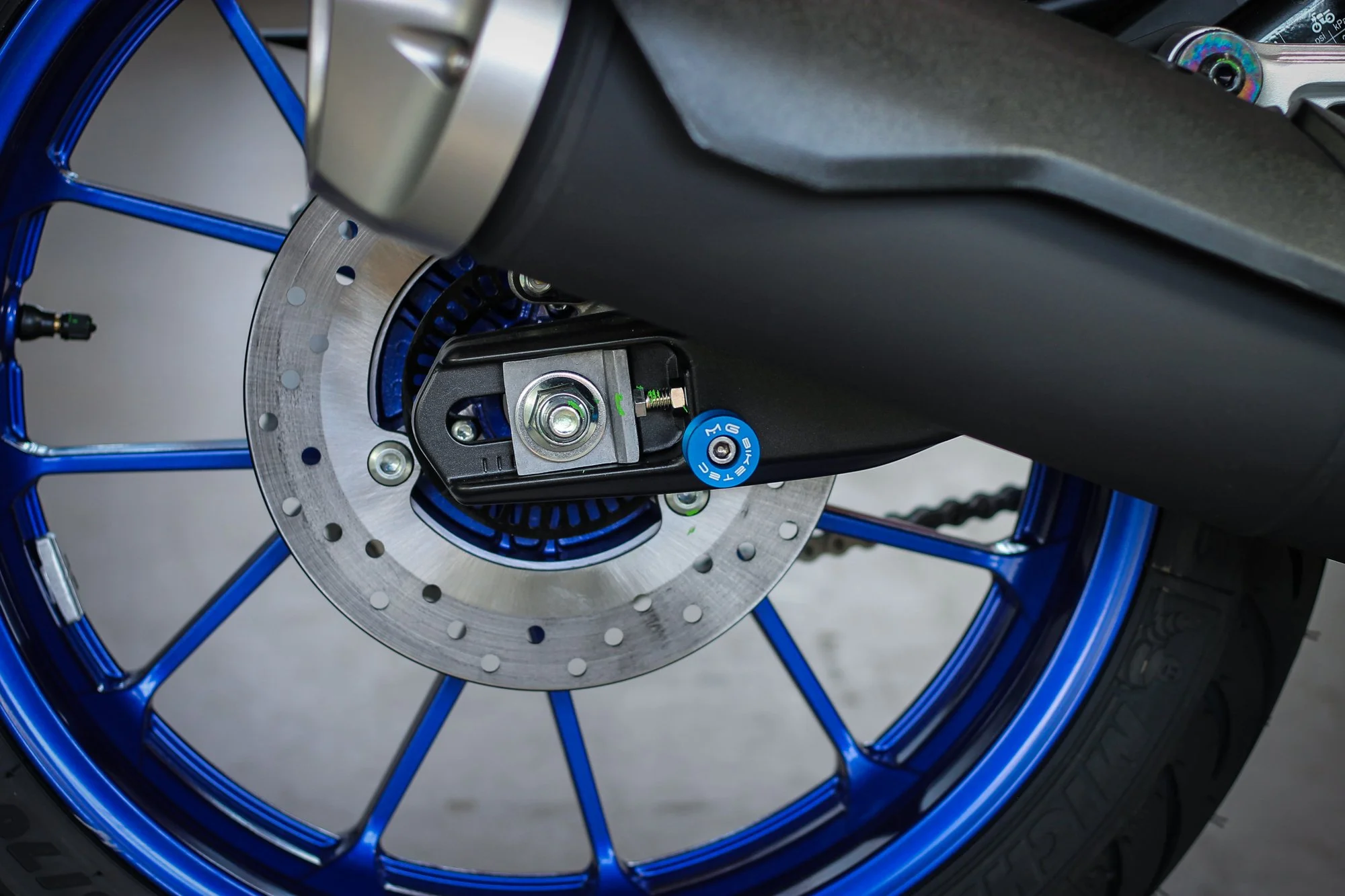 Close-up of a bicycle wheel featuring a disc brake rotor, blue spokes, and a black hub. A brake caliper and part of the frame are also visible.